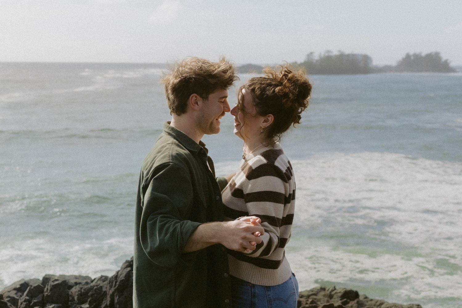 Happy couple holding hands and leaning in for a kiss on rocky coastal cliffs with ocean waves behind them.