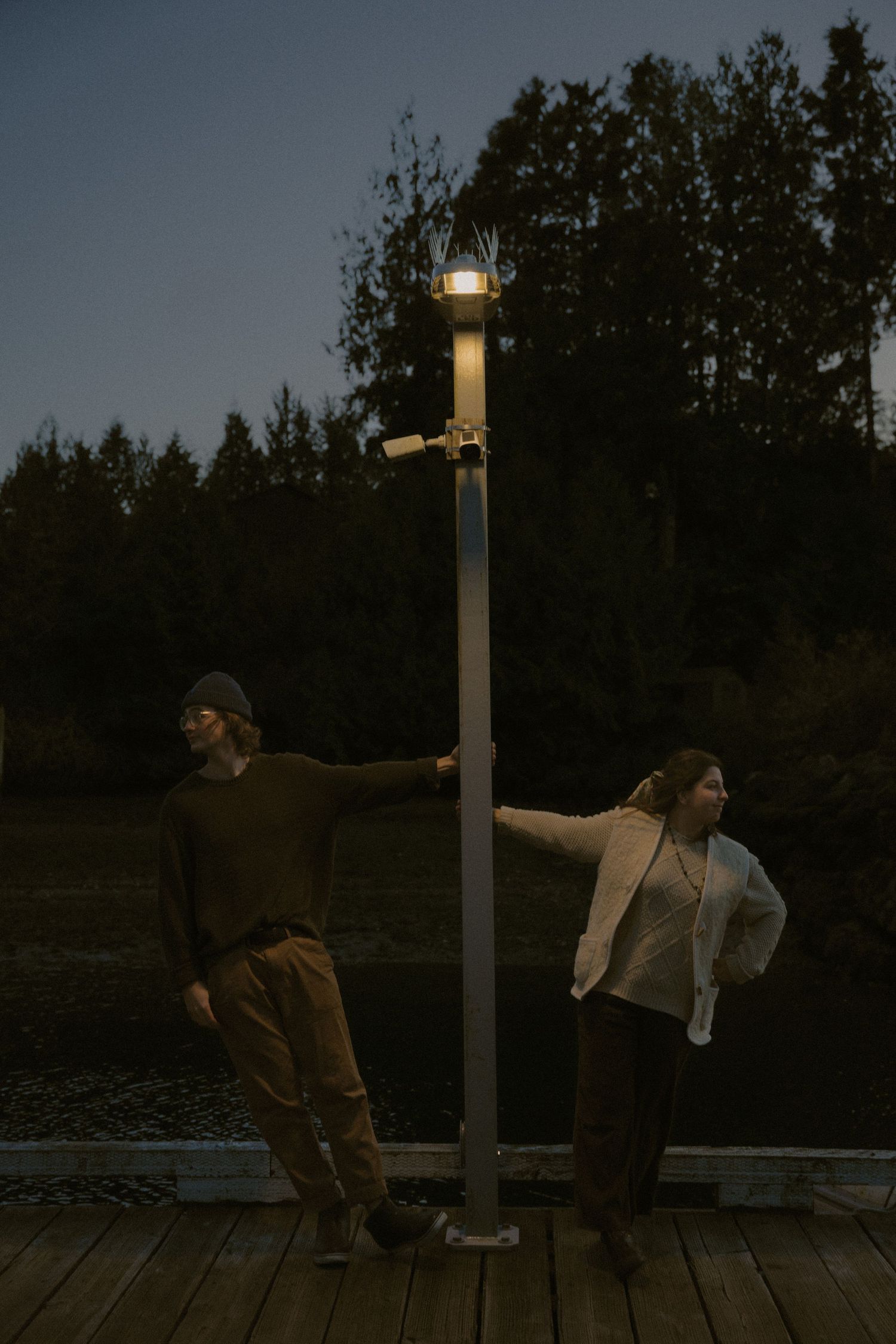 Two people dancing playfully on a wooden dock at twilight with lamp posts illuminating the scene.