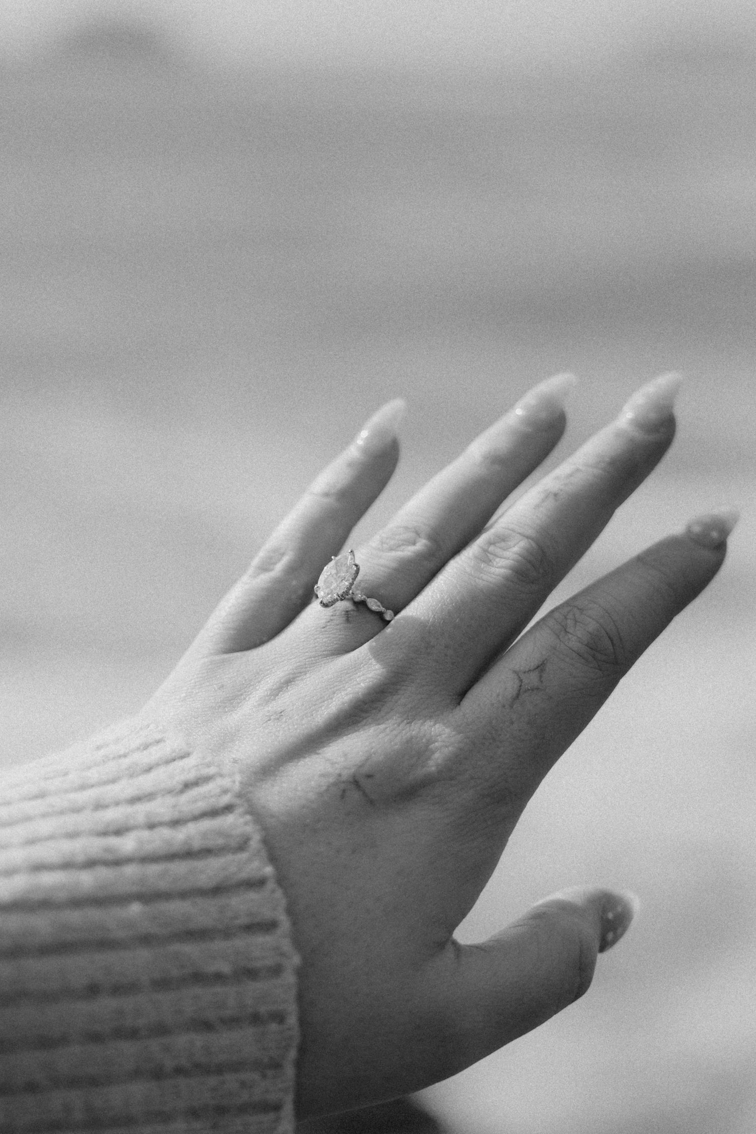 Close-up of a hand wearing an oval engagement ring, with almond-shaped nails and a cozy knit sweater sleeve.