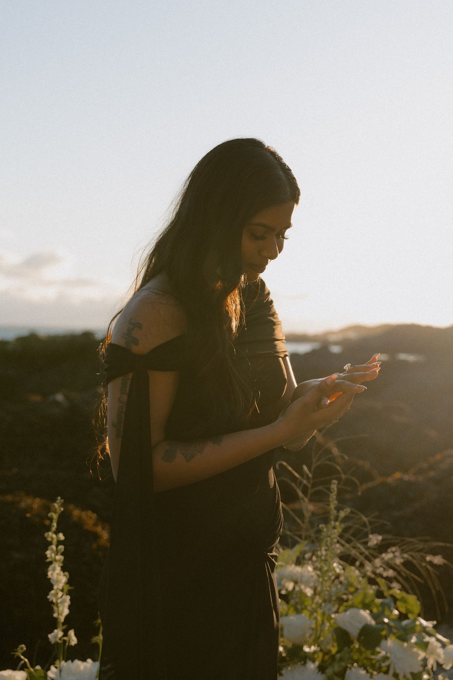 Young woman with long dark hair and tattoos examining a flower at sunset with scenic hills behind her.