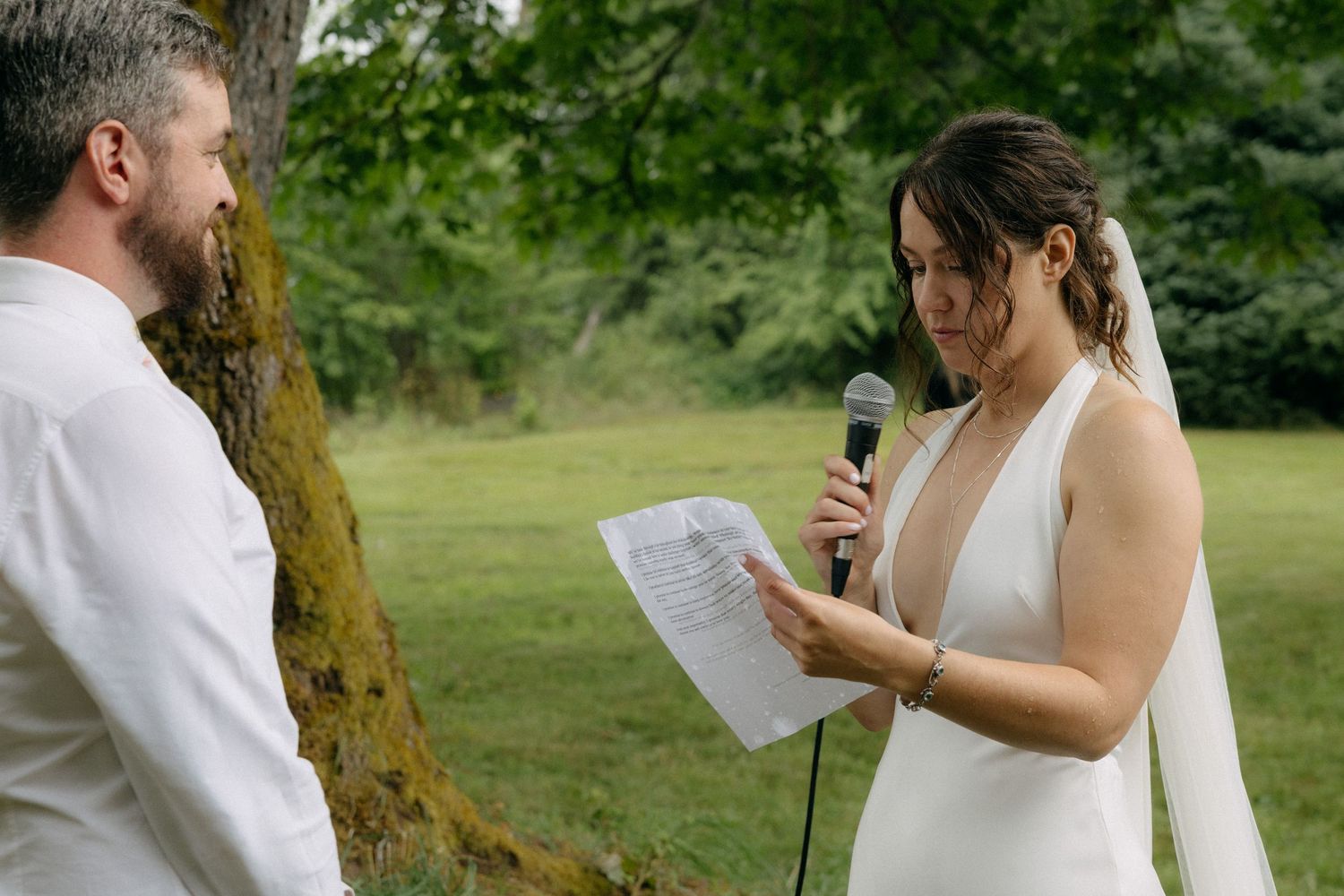 Bride reads wedding vows into microphone while groom listens outdoors near a mossy tree.