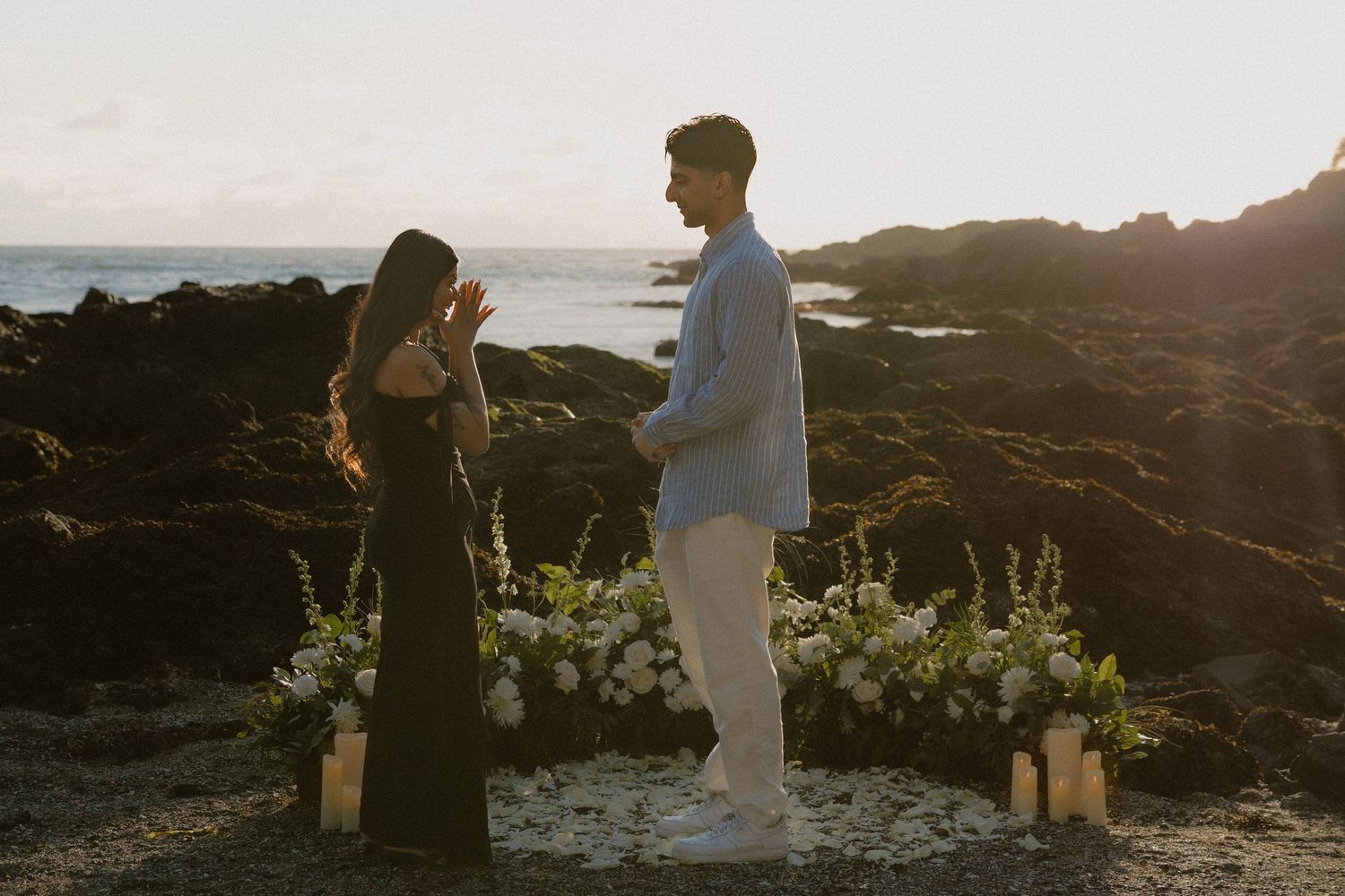Couple sharing a romantic beach proposal at sunset, surrounded by white flowers and candles on rocky Hawaiian coastline.