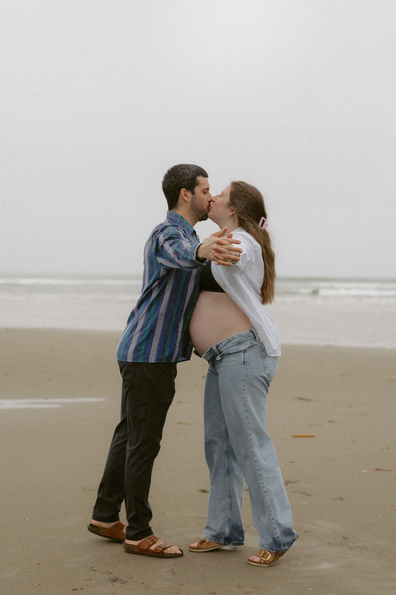 Two people share a tender kiss while standing on a misty beach in casual attire.