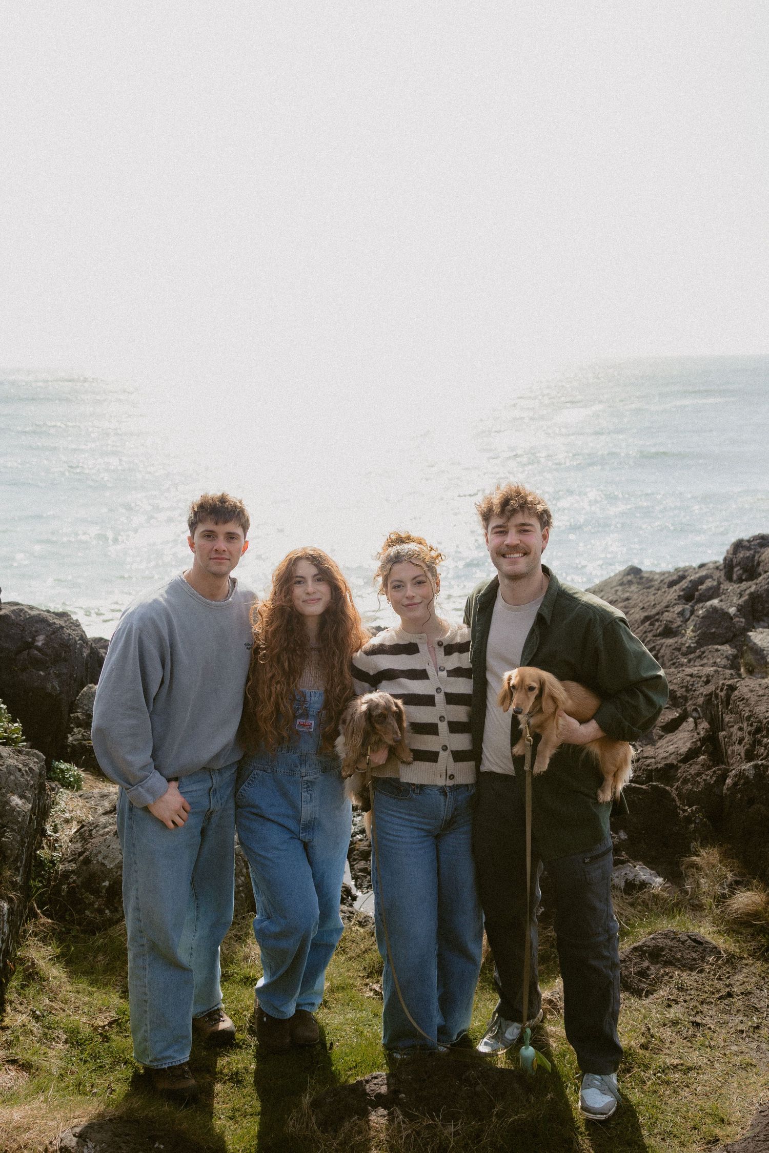 Four young friends with two golden dogs posing on rocky coastal cliffs with bright ocean backdrop.