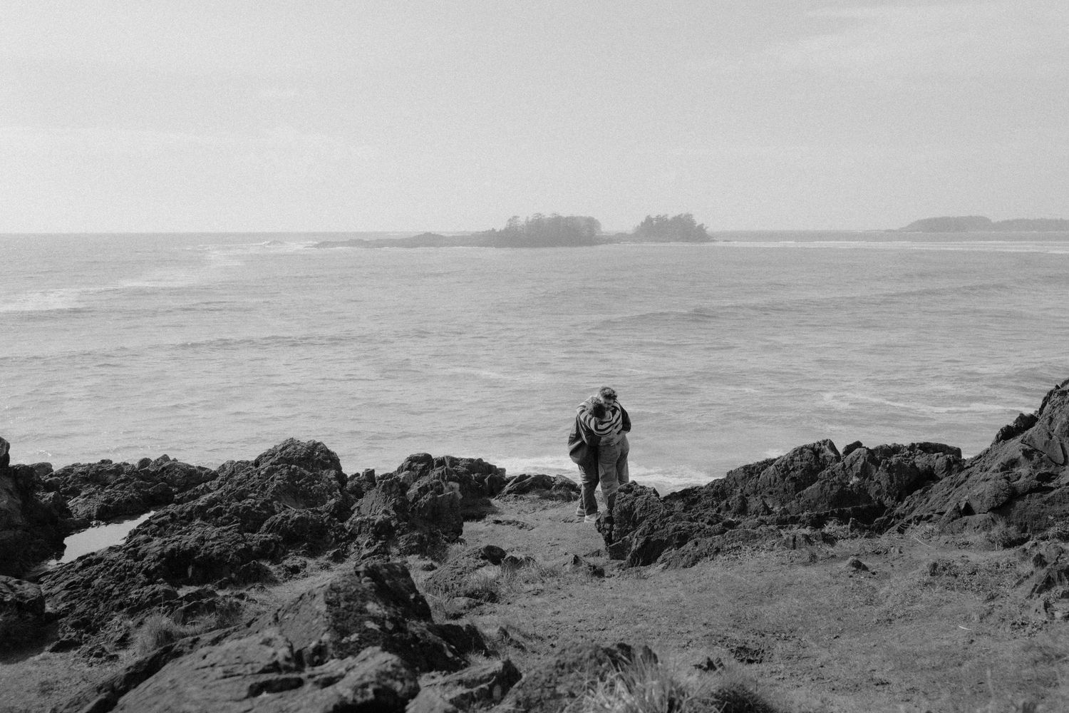 Person standing alone on rocky Pacific coastline in black and white photo with ocean waves.