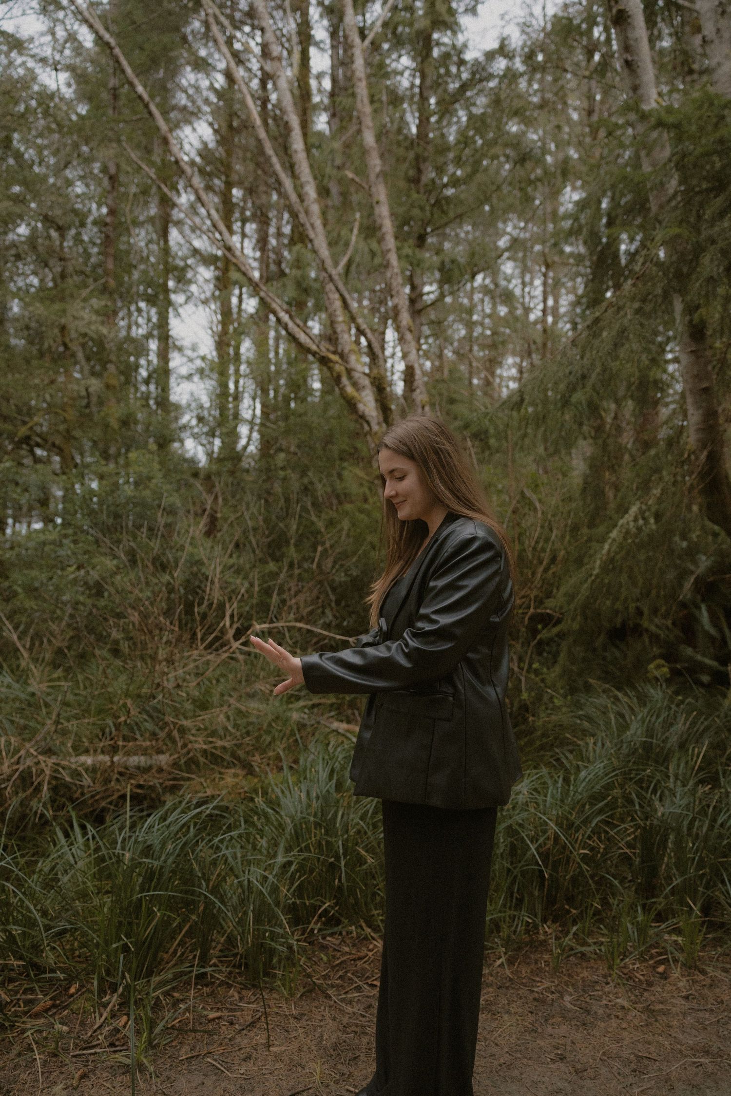 Person in black leather jacket checking phone while standing in wooded area on overcast day.
