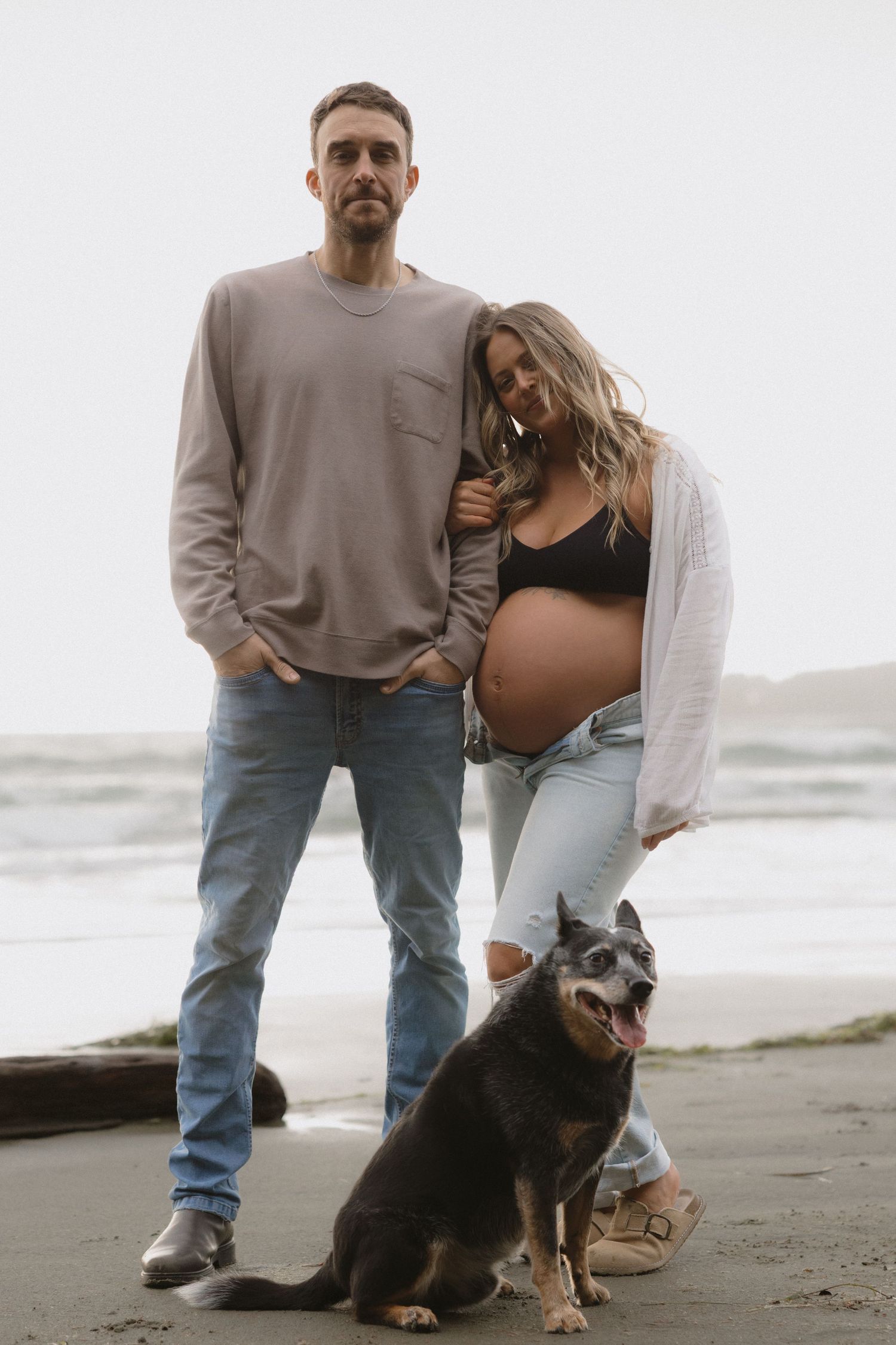 Couple with their dog poses for maternity photos on sandy beach at sunset.