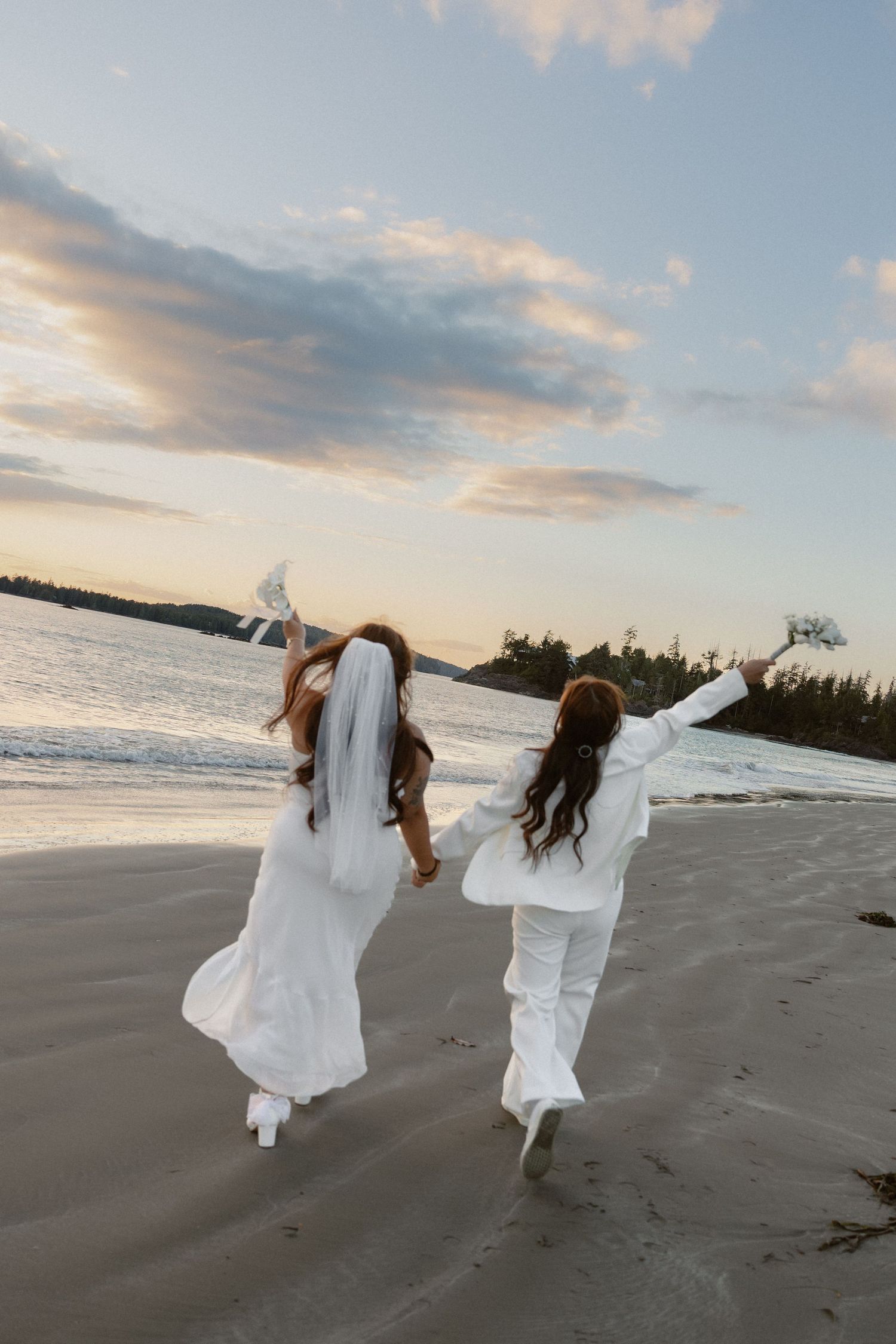 Two people in white celebrate with raised arms while walking on a beach at dusk.