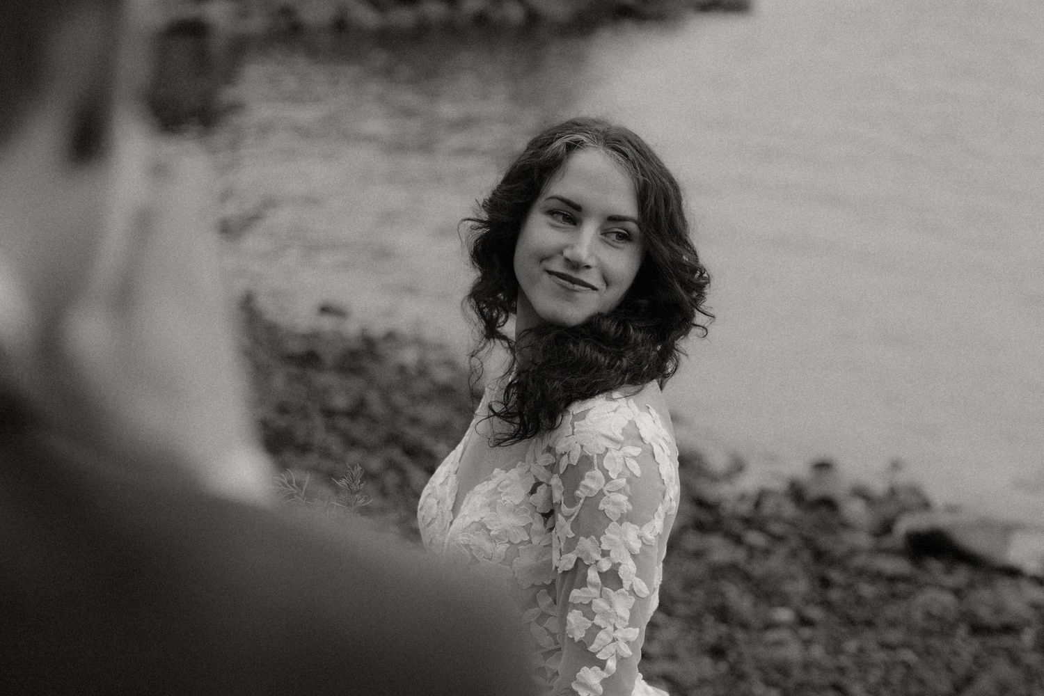 Black and white portrait of Tofino bride in lace dress looking over shoulder by the water