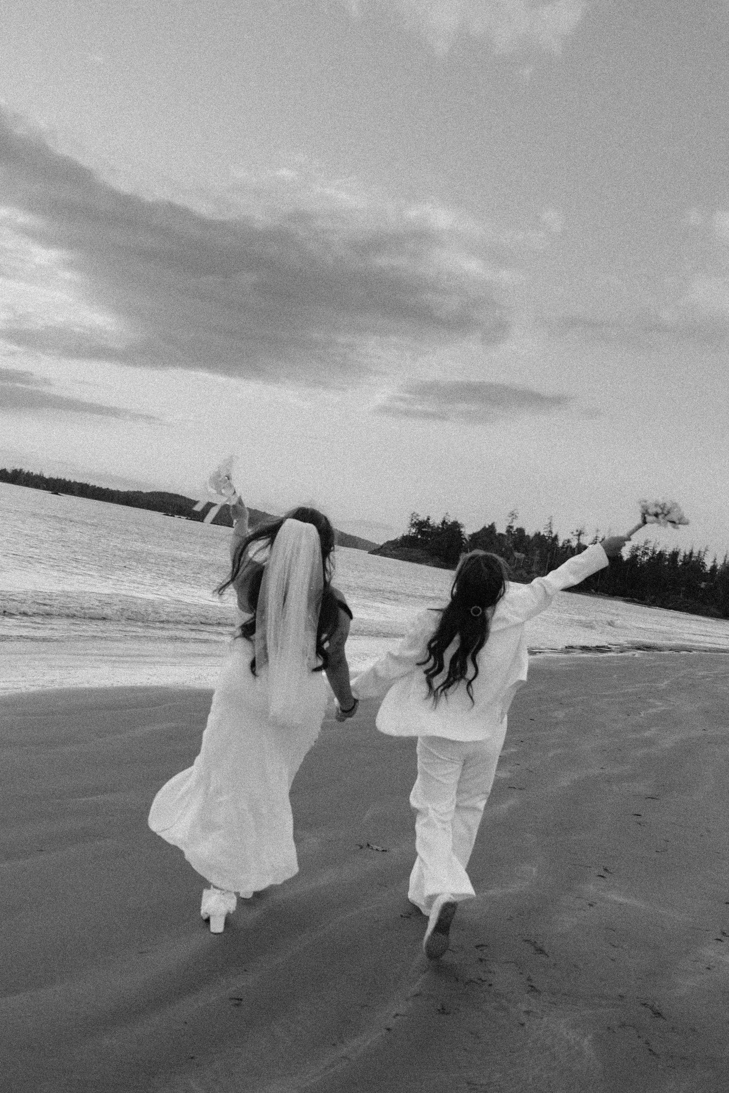 Two brides running down the beach in Tofino elopement, joyful moment with bouquet in the air