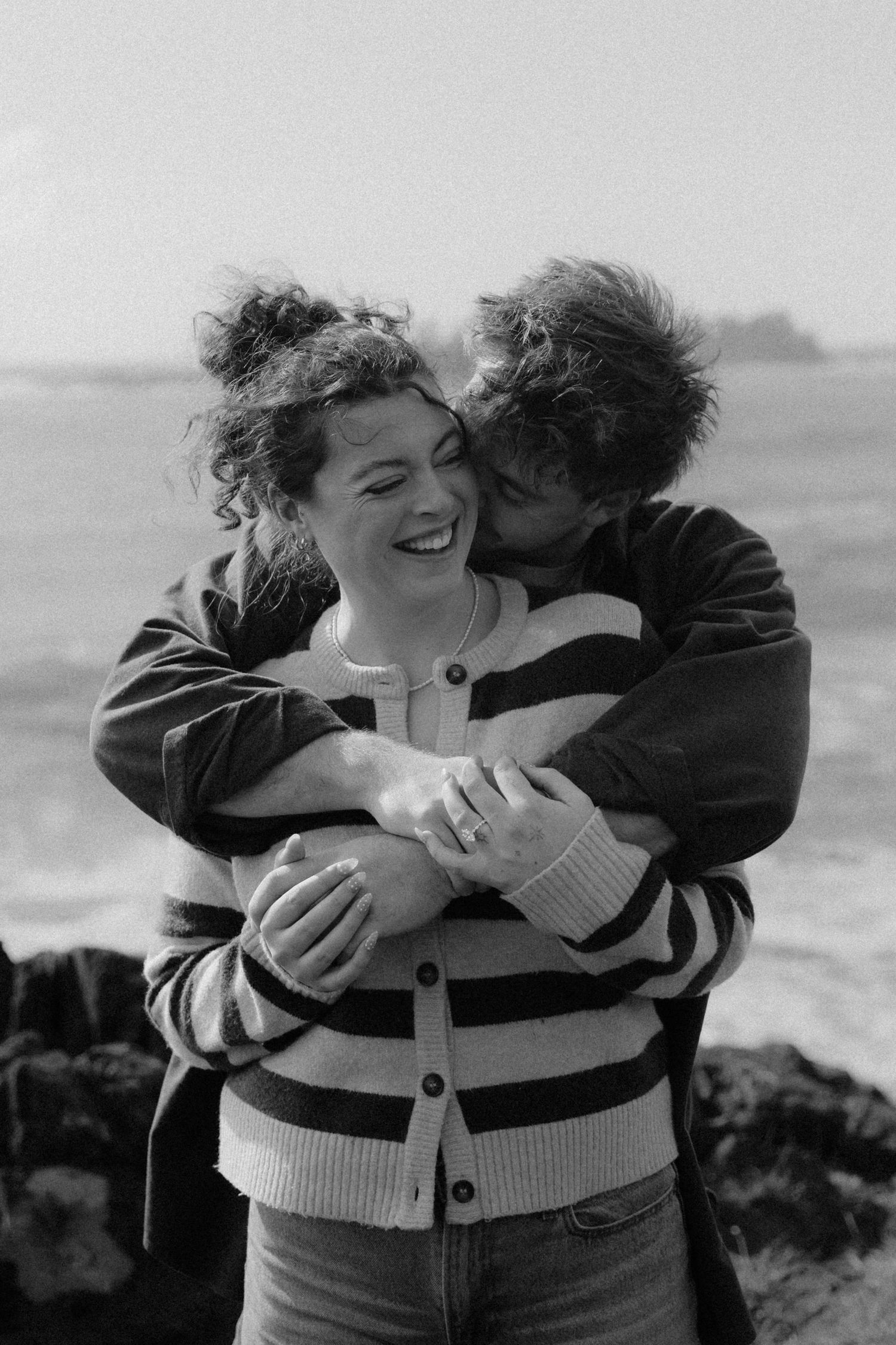Black and white photo of man embracing woman from behind as she laughs near ocean rocks.