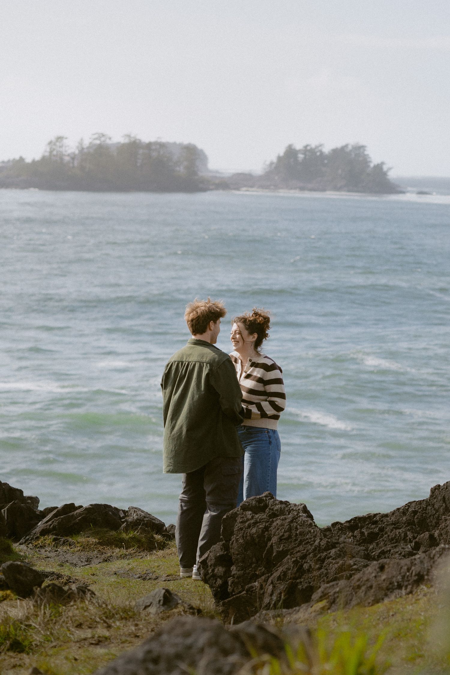 Couple standing on rocky coastal cliffs gazing at each other with ocean waves and island in background.