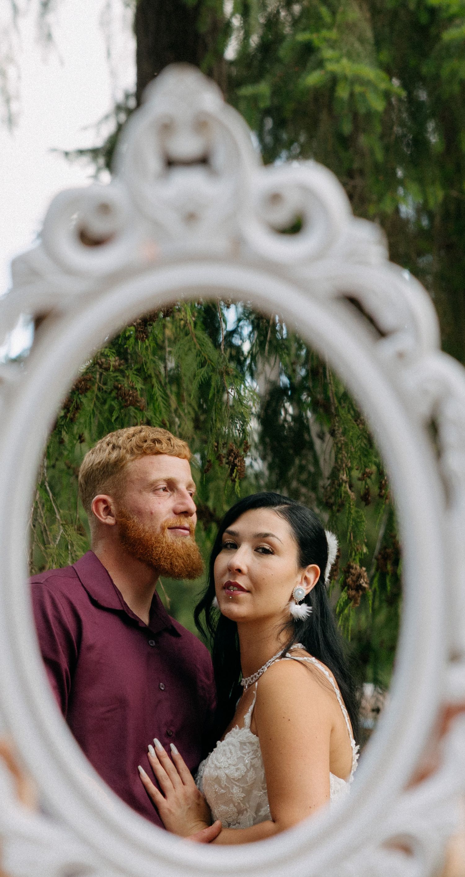 A couple poses romantically reflected in an ornate white oval mirror outdoors, bride in lace gown beside redbearded groom.