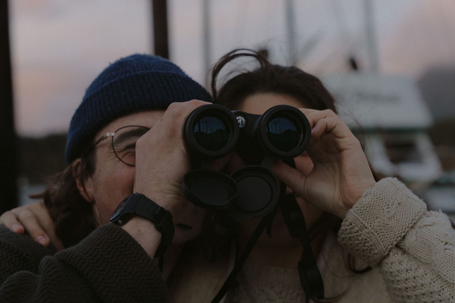 Tofino engagement couple share binoculars while standing at the Tofino harbor during golden hour