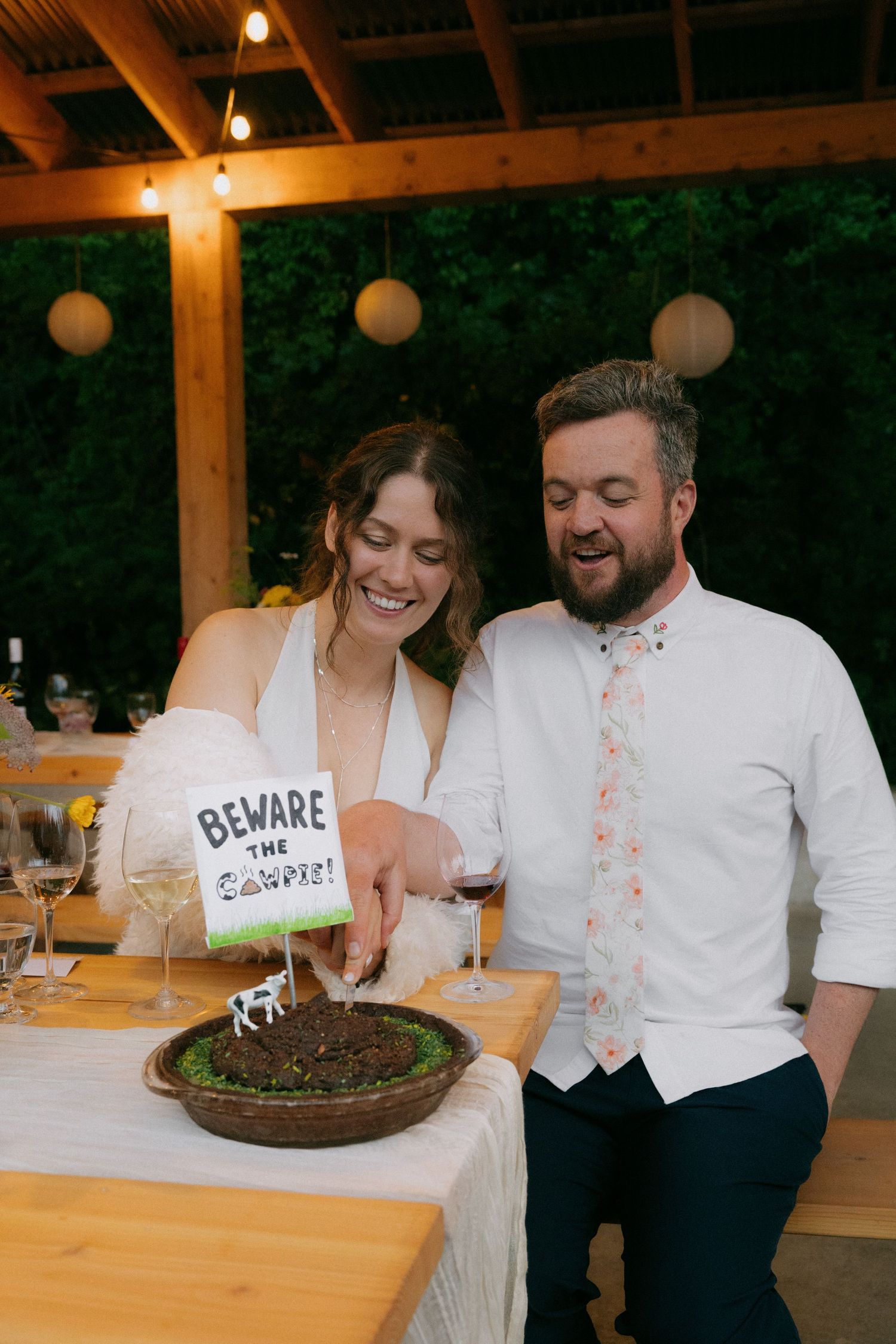 Laughing bride and groom cut a humorous dirt cake with a Beware the Cows sign at reception.