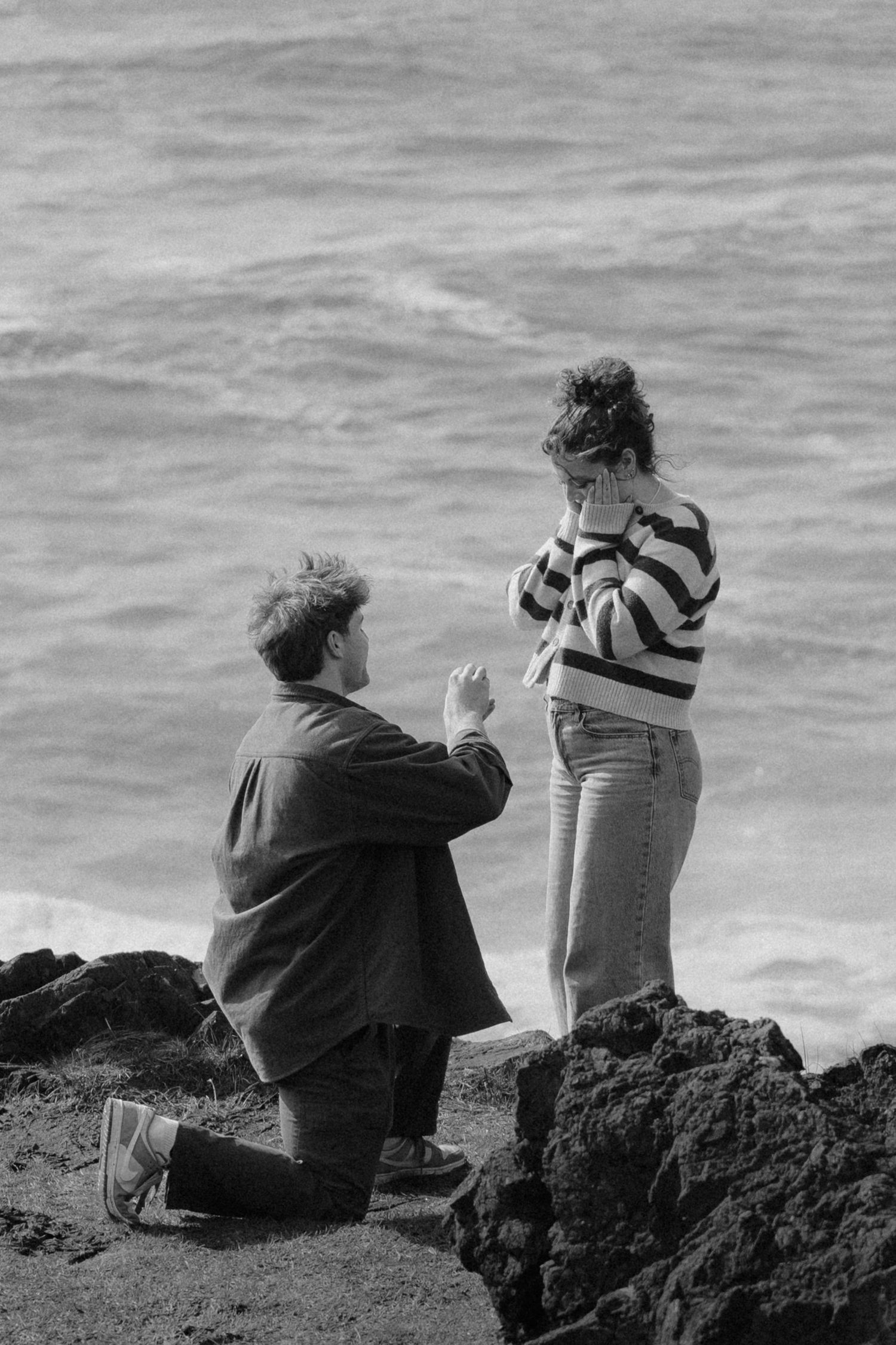 Man proposing on one knee to emotional woman on rocky seaside cliffs in black and white.