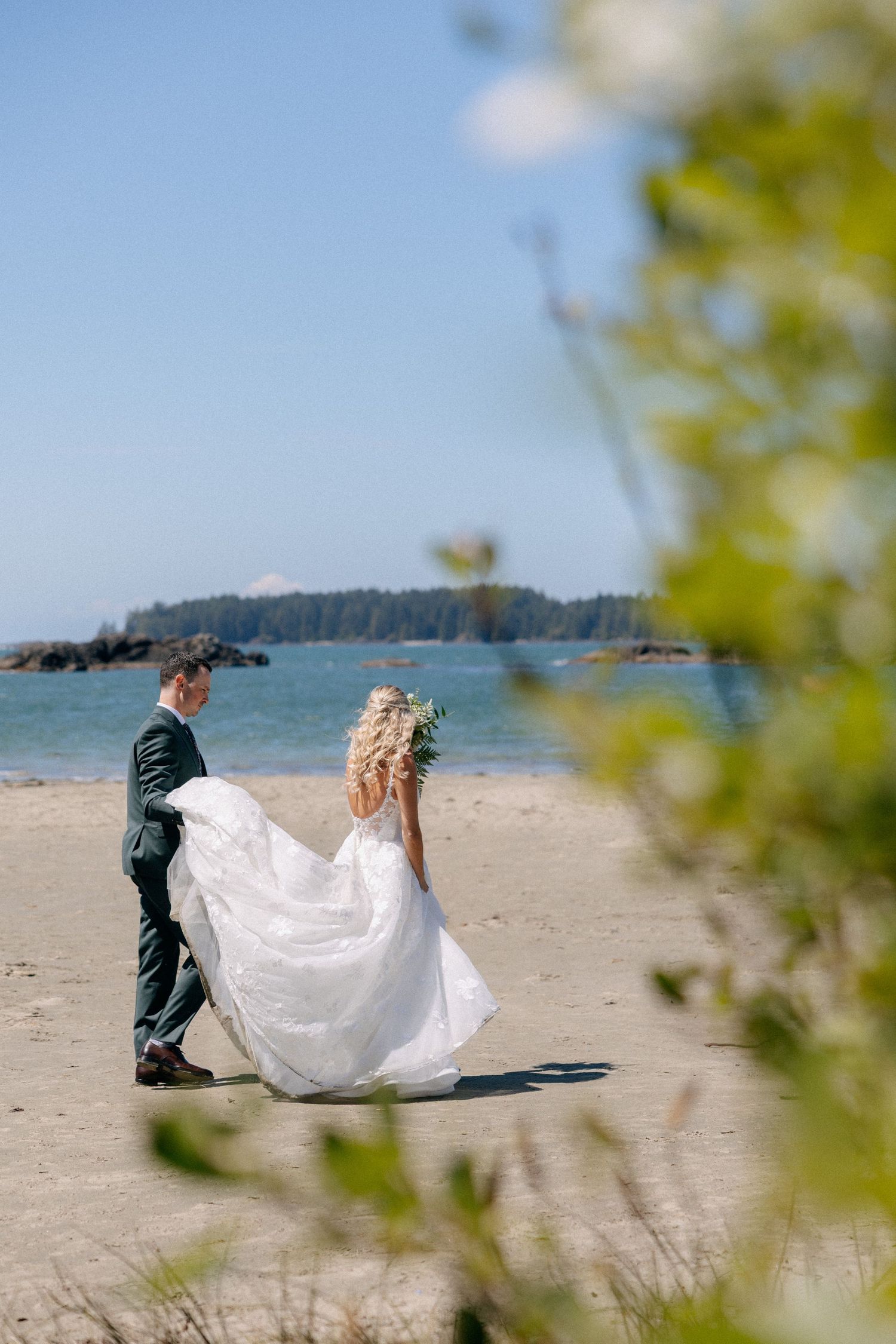 Bride and groom walking on a sandy beach in front of blue ocean water, bride's white gown flowing in the breeze.