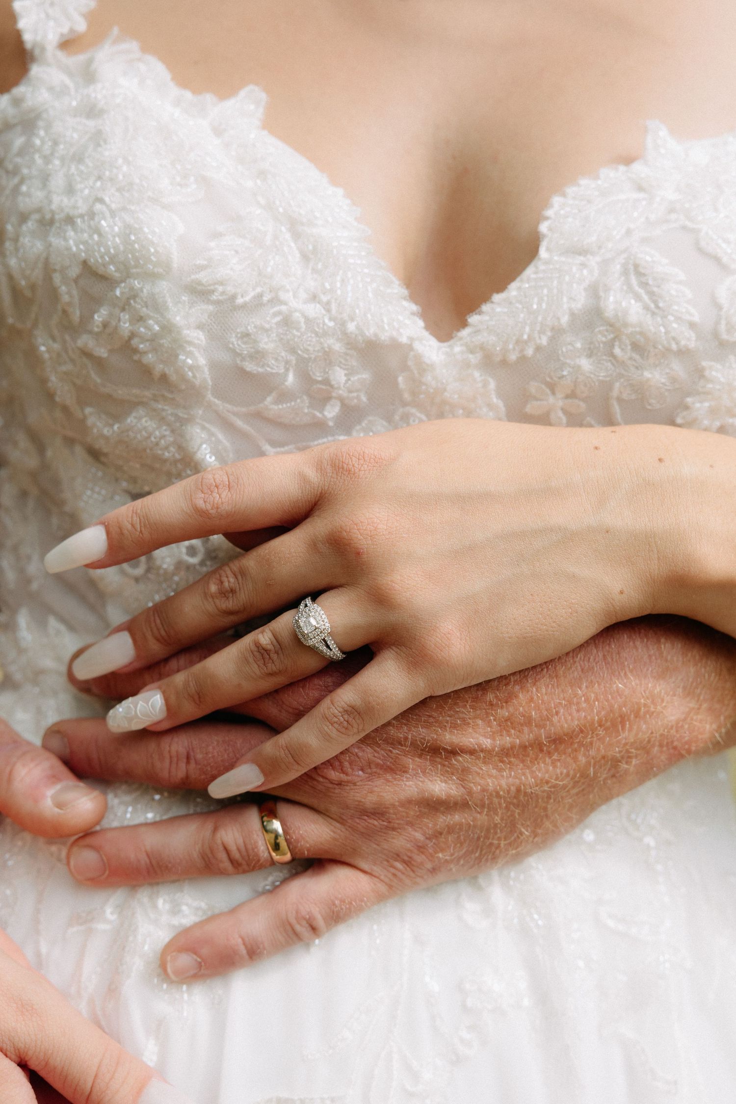 Close-up of bride and groom hands showing diamond engagement ring and gold wedding band on white lace dress.