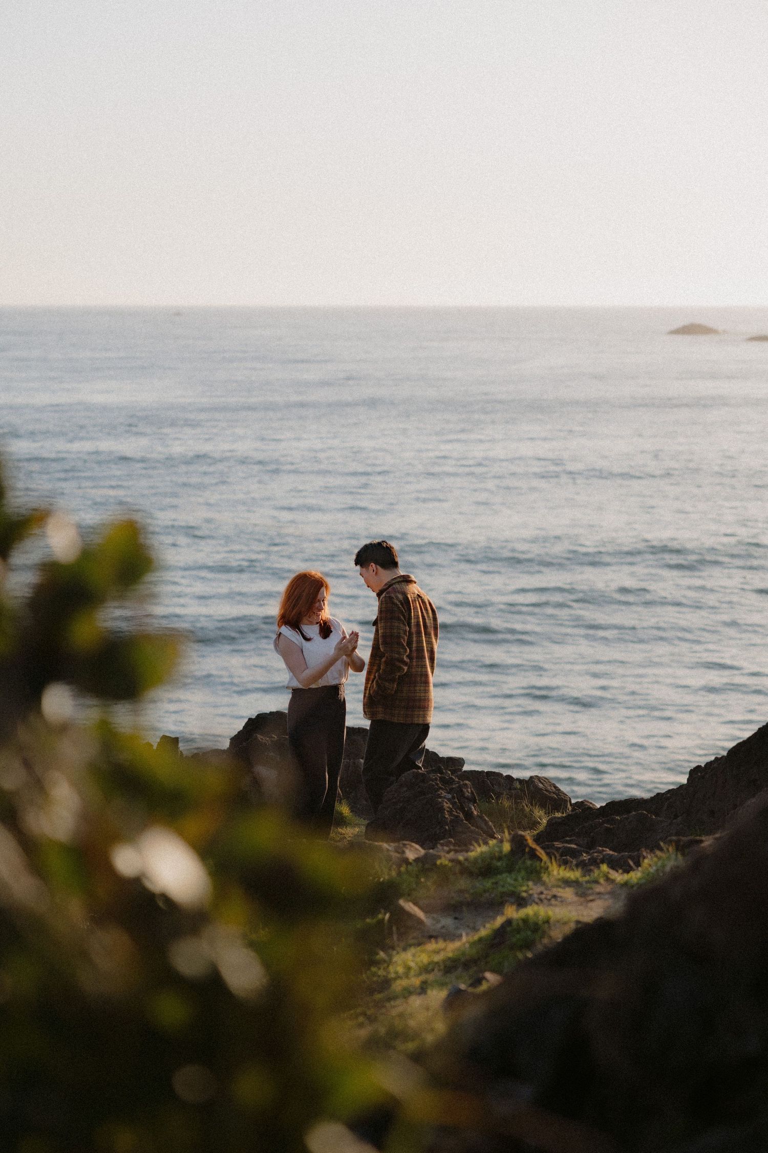 Couple standing on rocky coastal cliffs with ocean backdrop during golden hour engagement photo session.
