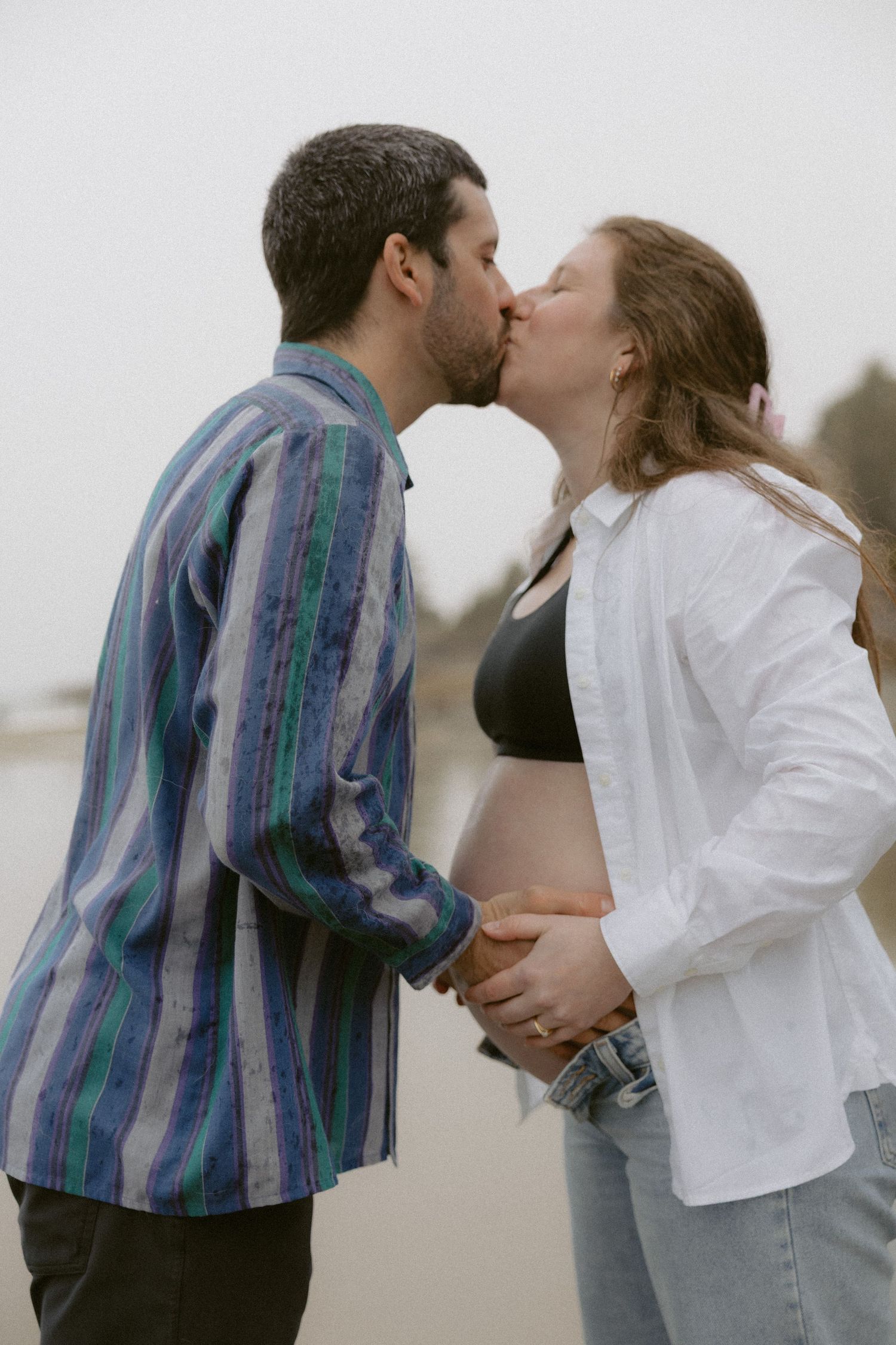 A couple shares a tender kiss on a cloudy beach, with the woman wearing a white jacket and the man in a striped shirt.