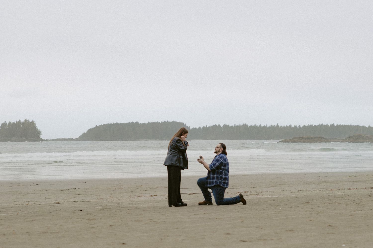 Two figures stand on a foggy beach with distant coastal cliffs visible through the mist.