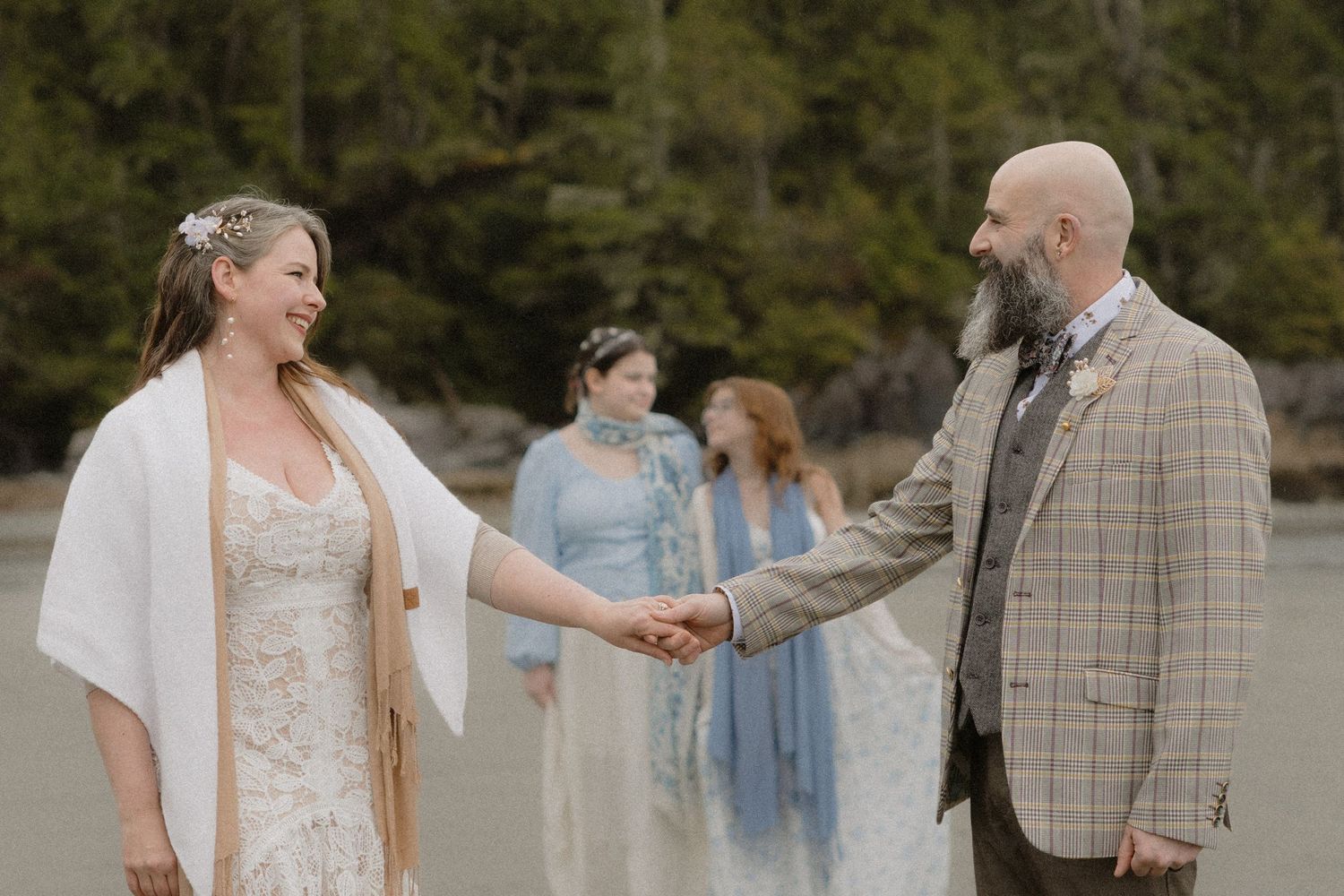 Couple holds hands during beach wedding ceremony with attendants in blue dresses watching in background.