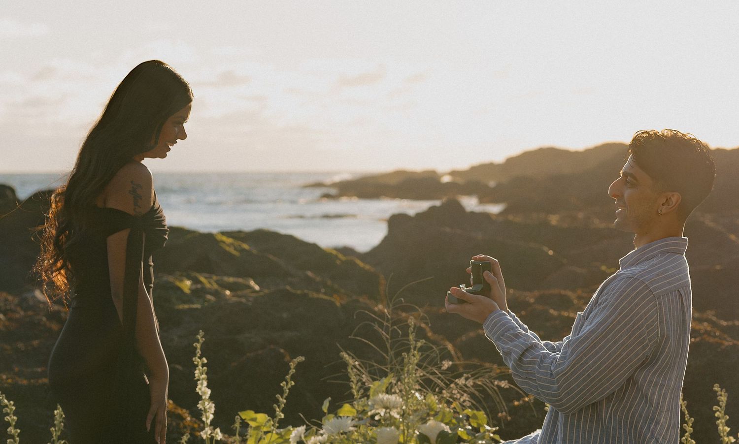 Man proposes to woman on rocky coastal cliff at sunset, holding engagement ring while kneeling outdoors.