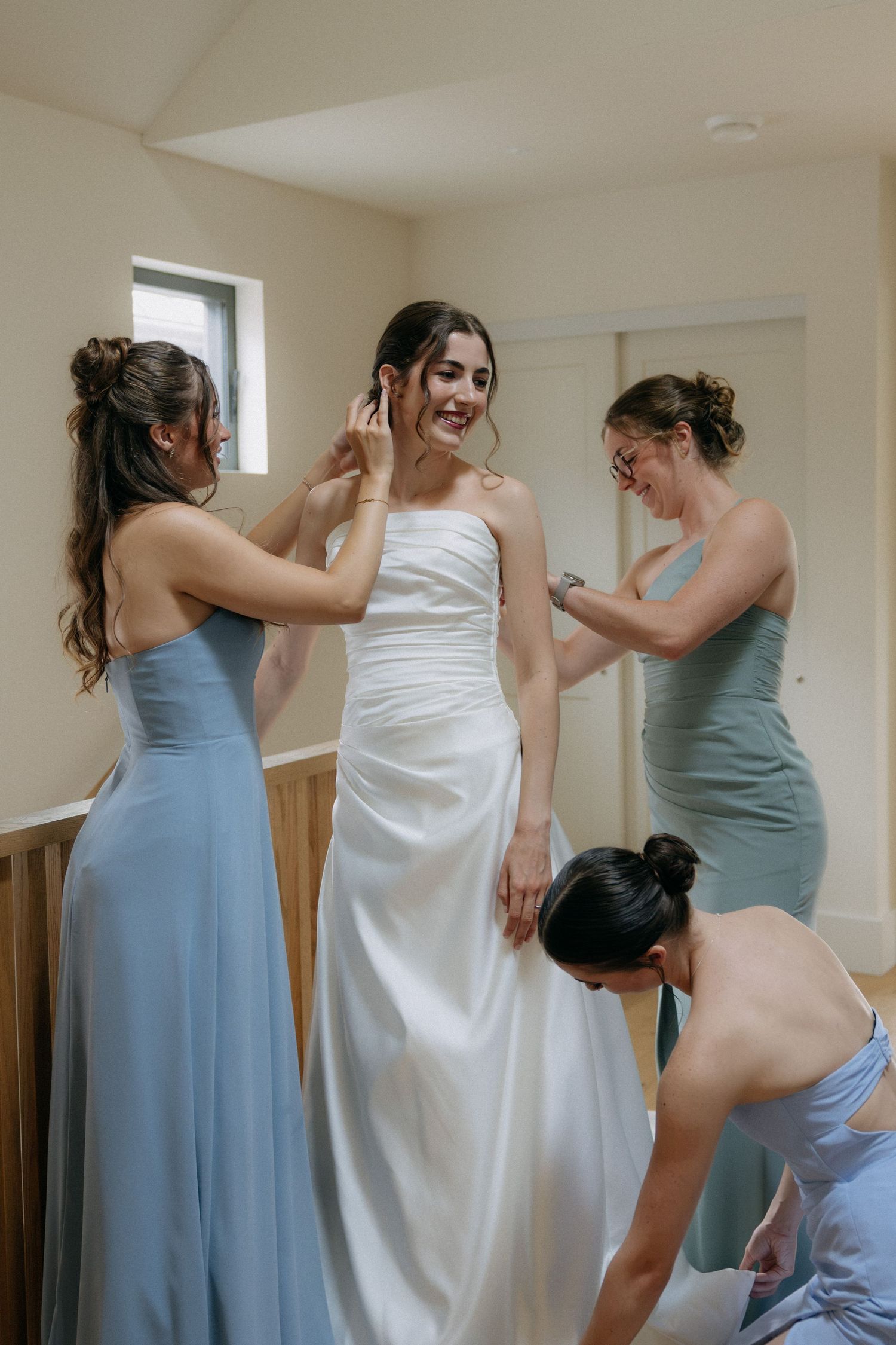 Bride smiling as three bridesmaids in blue dresses help her get ready in a bright room.