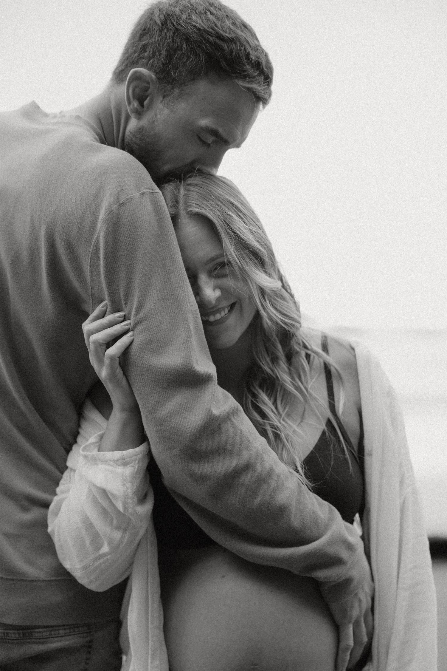 Black and white intimate maternity portrait showing couple embracing on beach.
