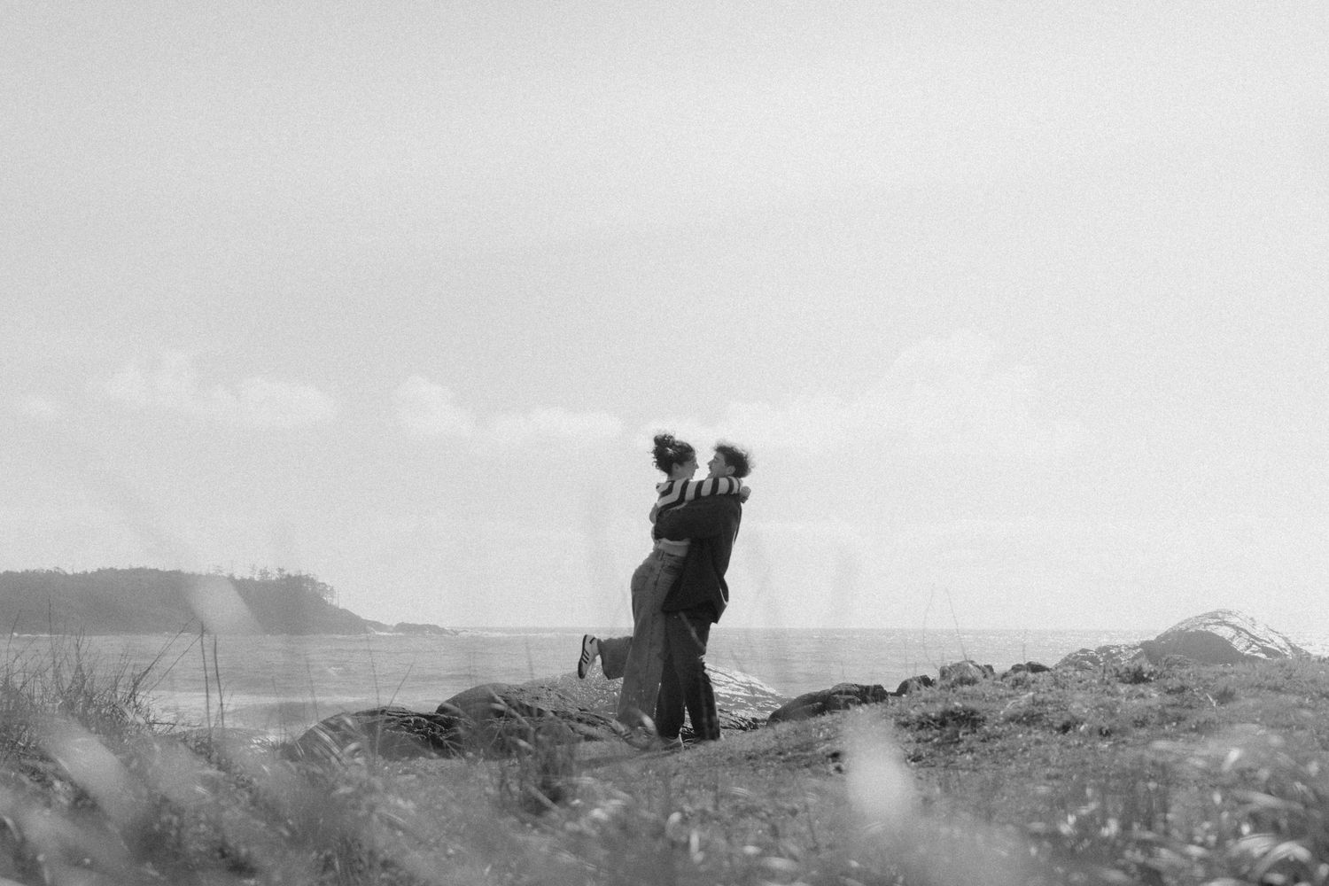 Black and white photo of a romantic couple embracing on a rugged coastal landscape with the ocean behind them.