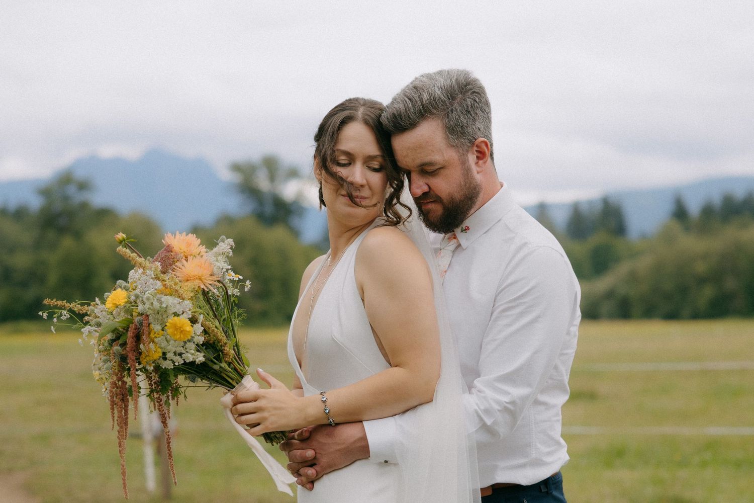 Couple embracing in a meadow with mountain backdrop, bride holding wildflower bouquet in yellow and pink hues.