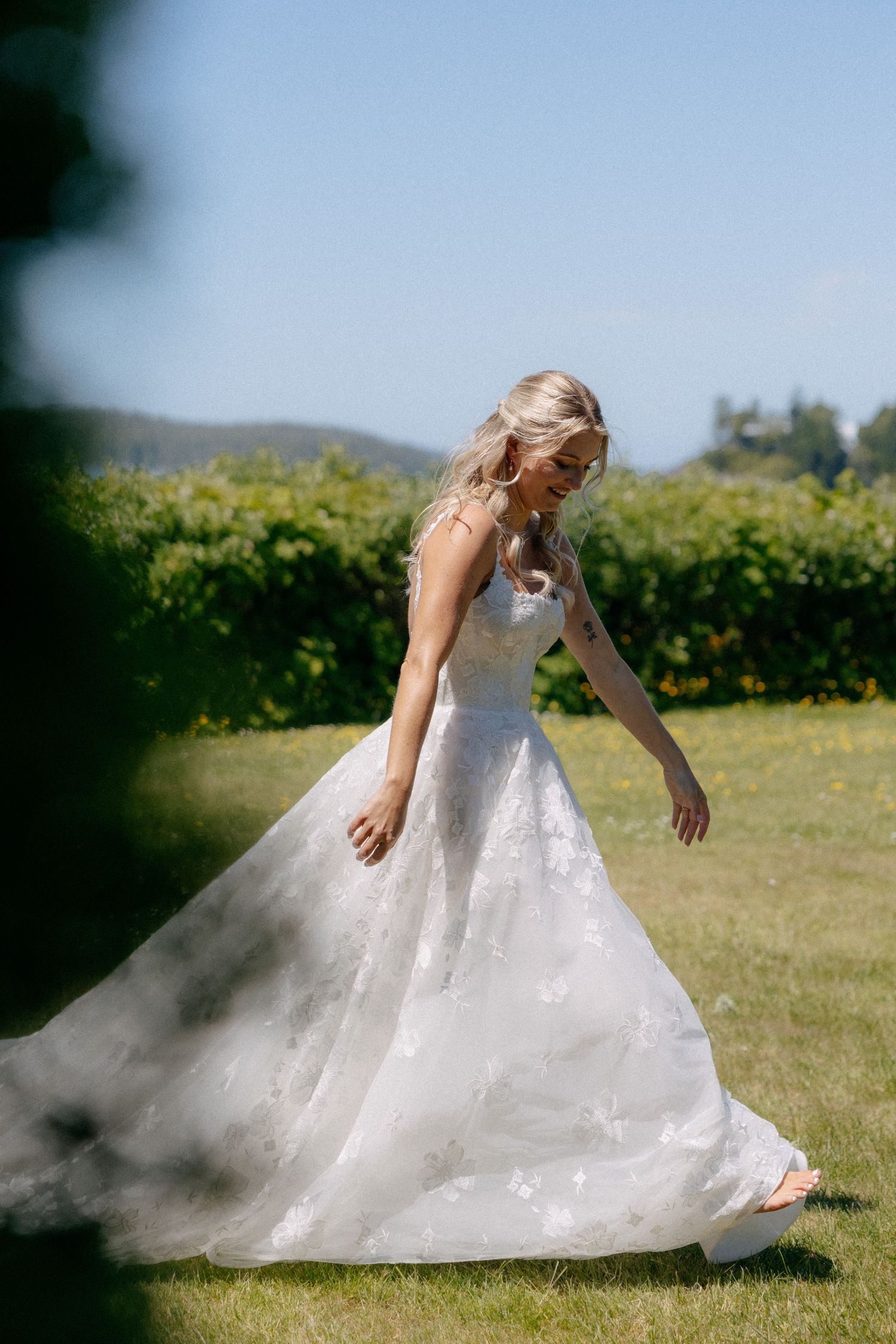 Bride in a flowing white floral wedding gown walks barefoot on green grass outdoors on a sunny day.