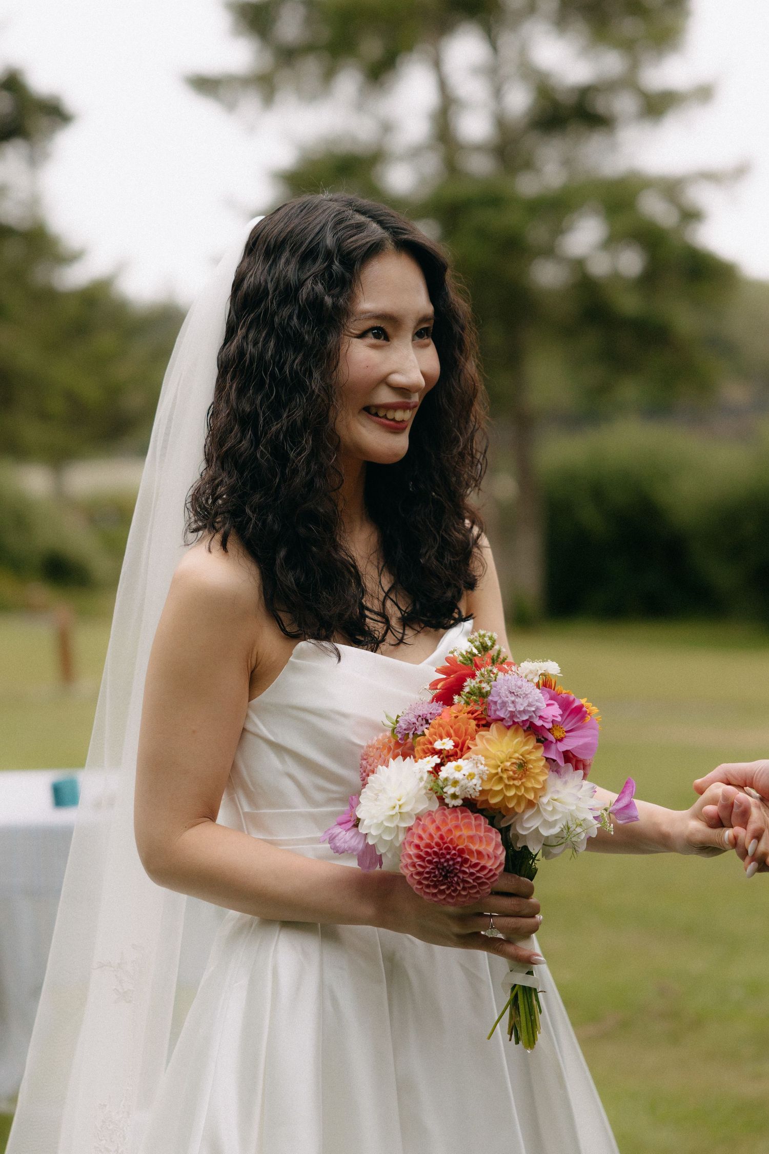 Smiling Asian bride in white strapless gown and veil holds colorful dahlia bouquet outdoors at garden wedding.