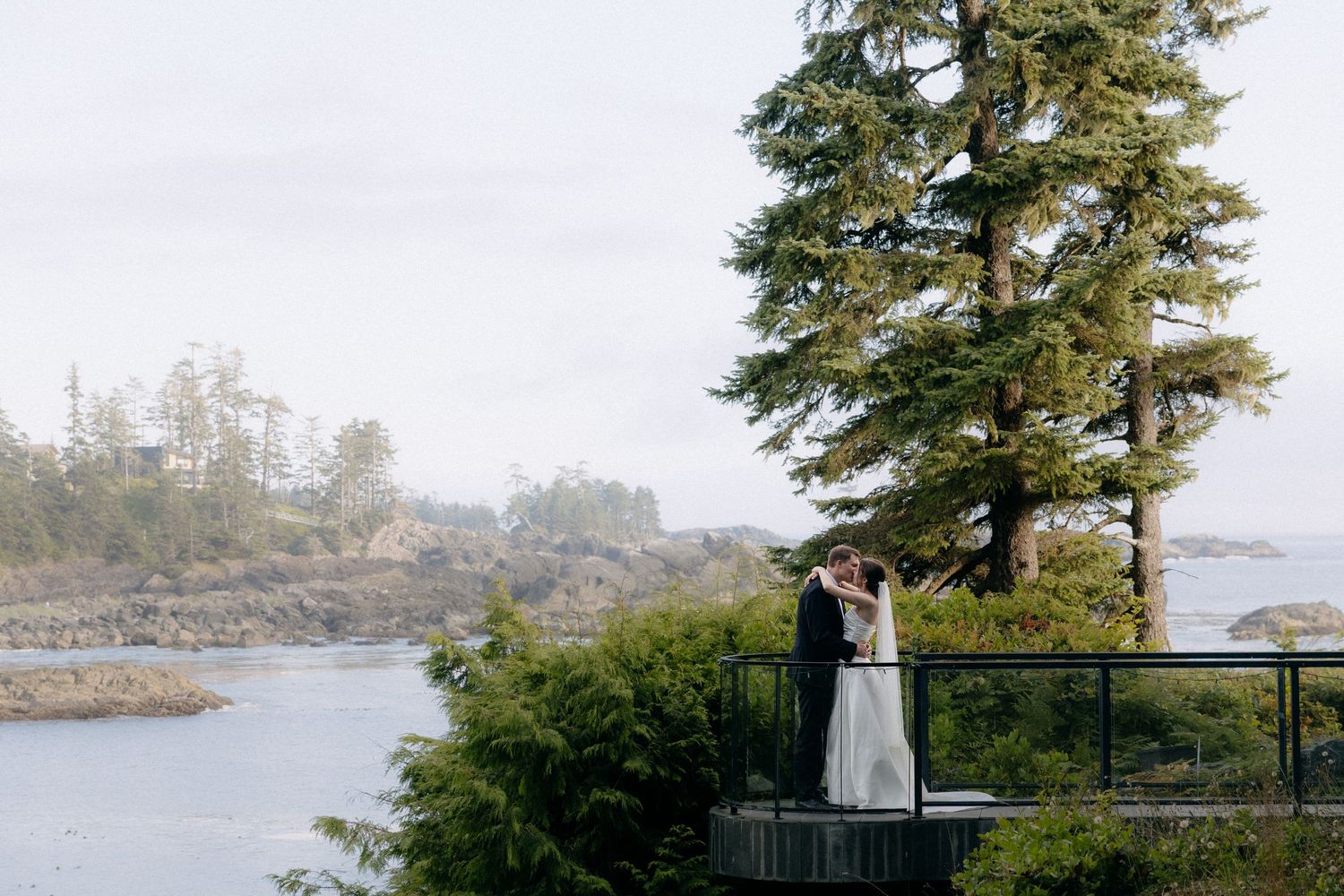 Bride and groom share a kiss on a scenic overlook deck surrounded by tall trees and ocean views on their wedding day.