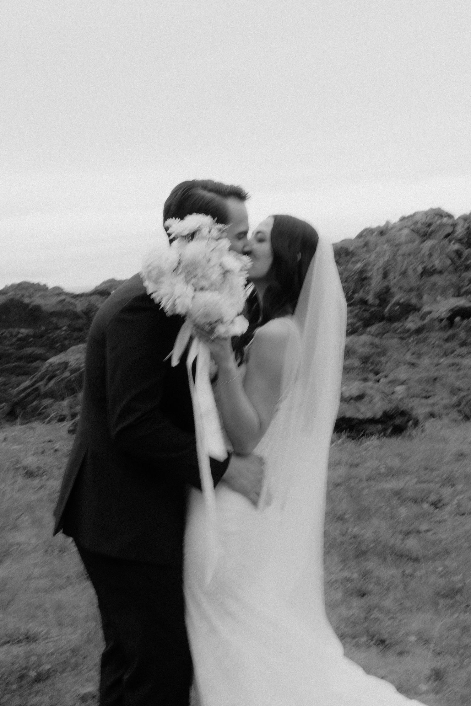 Black and white photo of a bride and groom sharing a kiss outdoors, bride holding a bouquet with veil flowing.