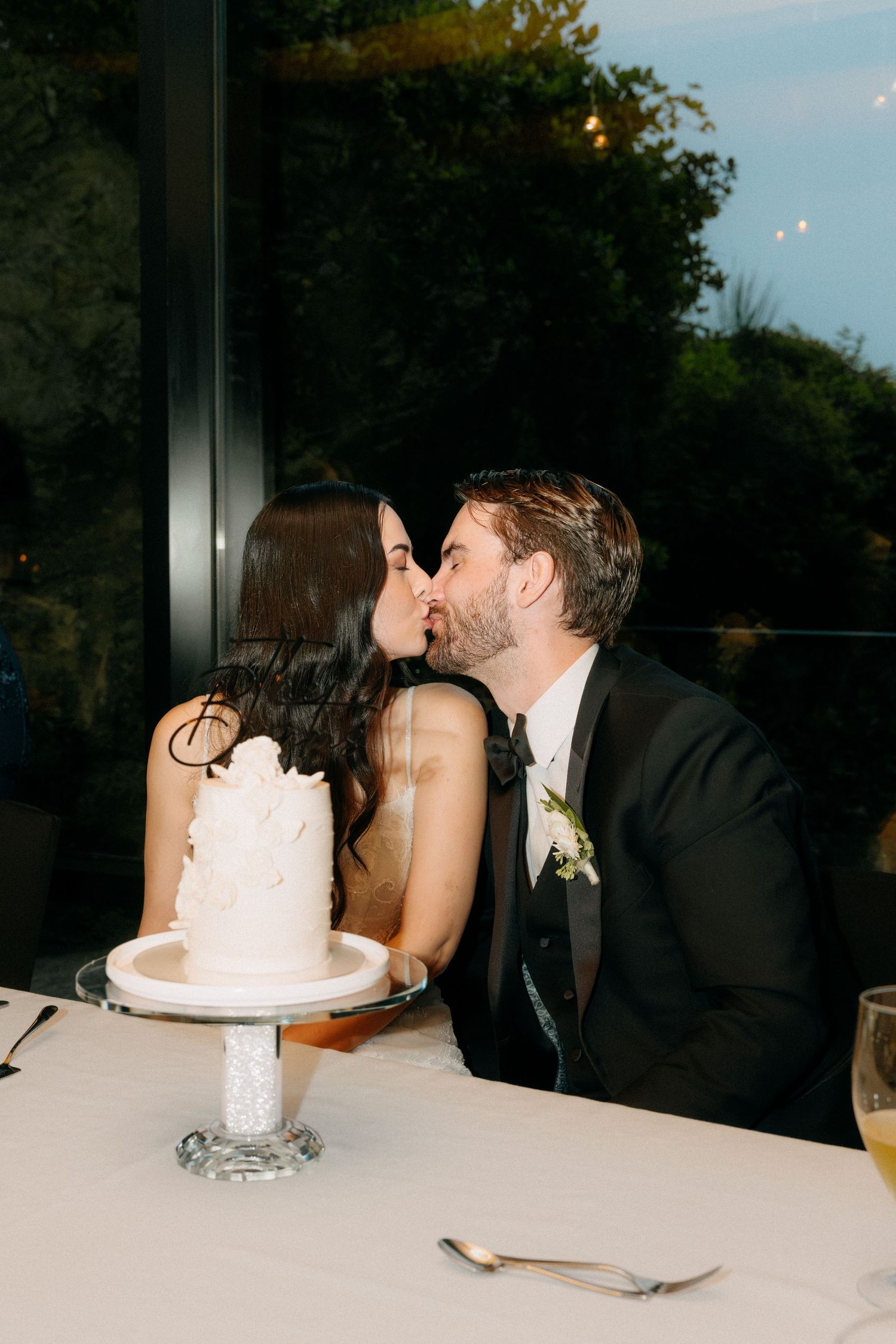 Bride and groom sharing a kiss at wedding reception table with white tiered cake on crystal stand.
