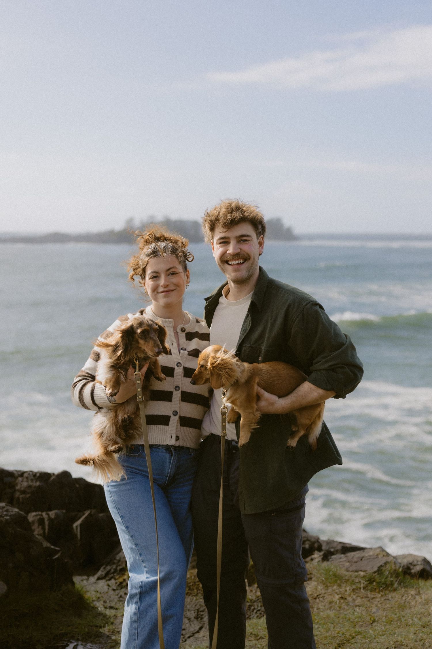 Smiling couple holding two golden dogs on a rocky coastline with blue ocean and sky backdrop.