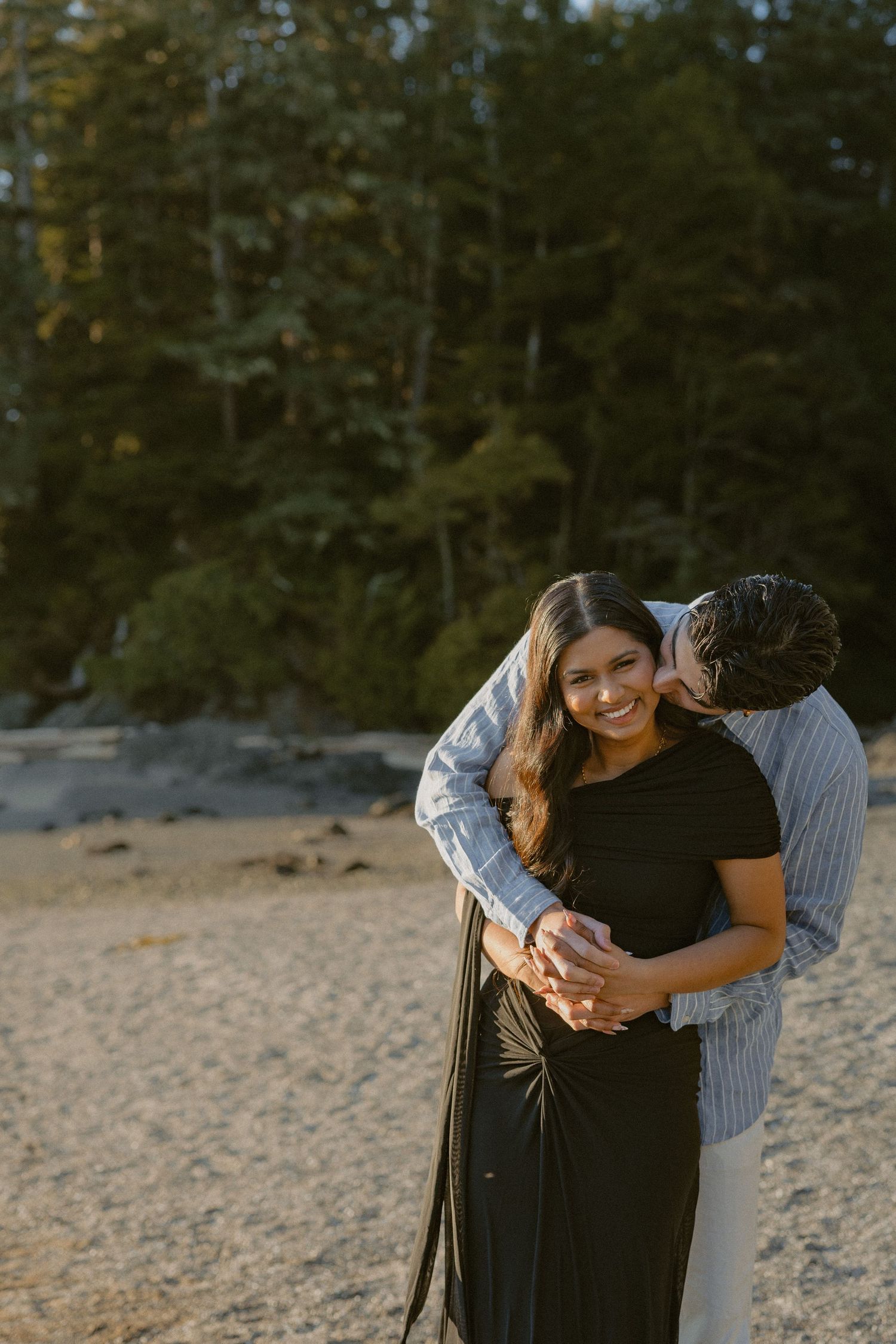Couple embracing on sandy beach at golden hour, woman smiling as man kisses her cheek near forest backdrop.