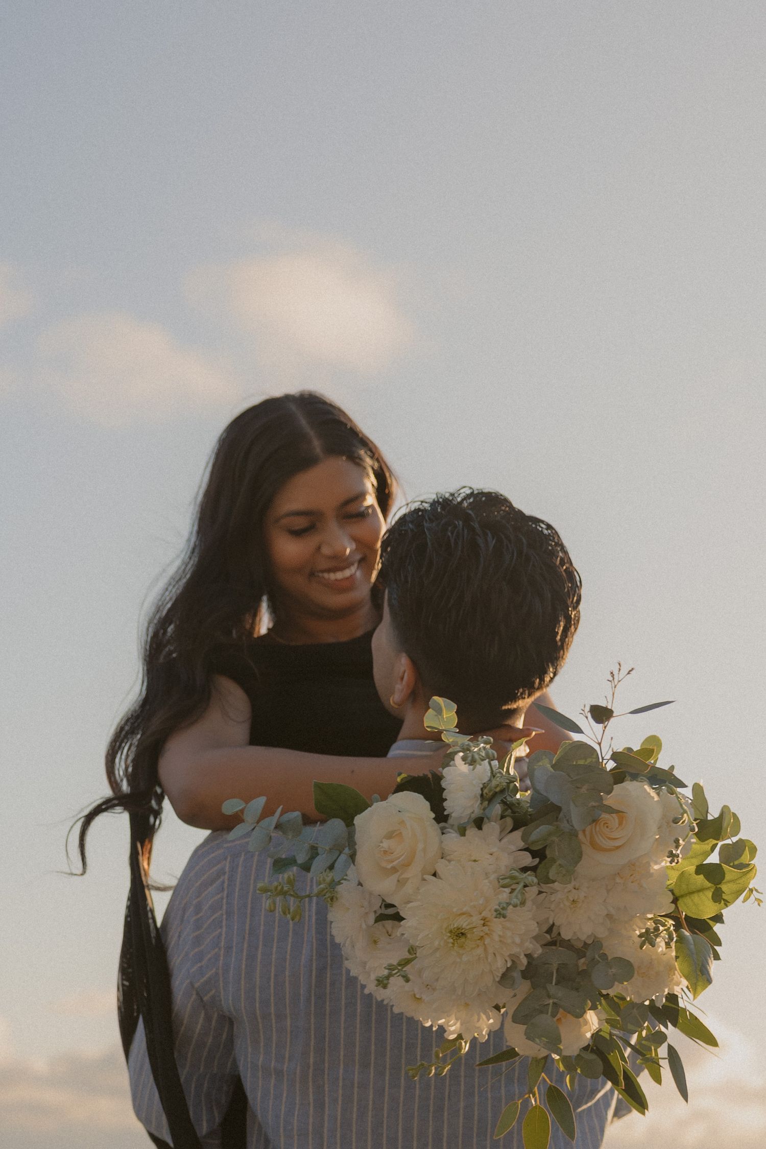 Smiling woman embracing man holding a bouquet of white roses and greenery at sunset.