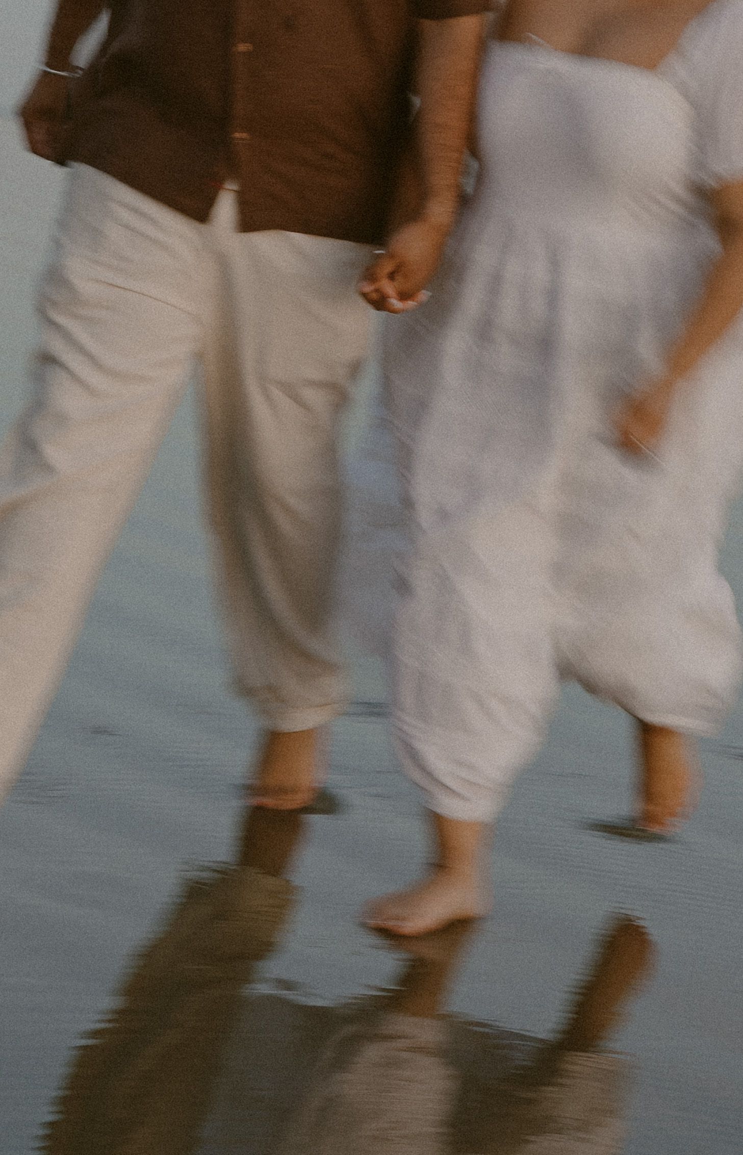 Couple holding hands walking barefoot on a wet beach, wearing white linen outfits, captured in a dreamy motion blur.