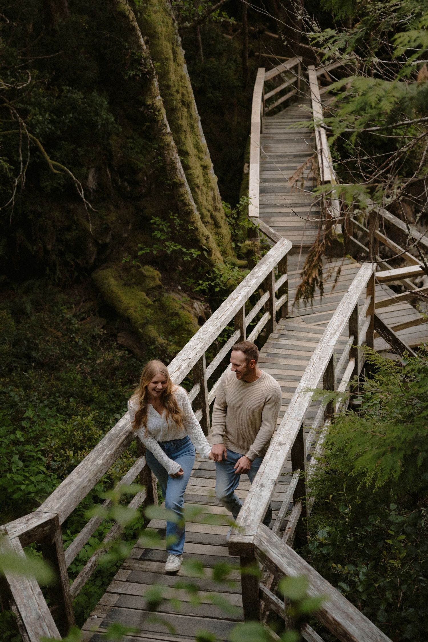Couple holding hands walking down wooden forest stairs surrounded by lush green moss-covered trees.