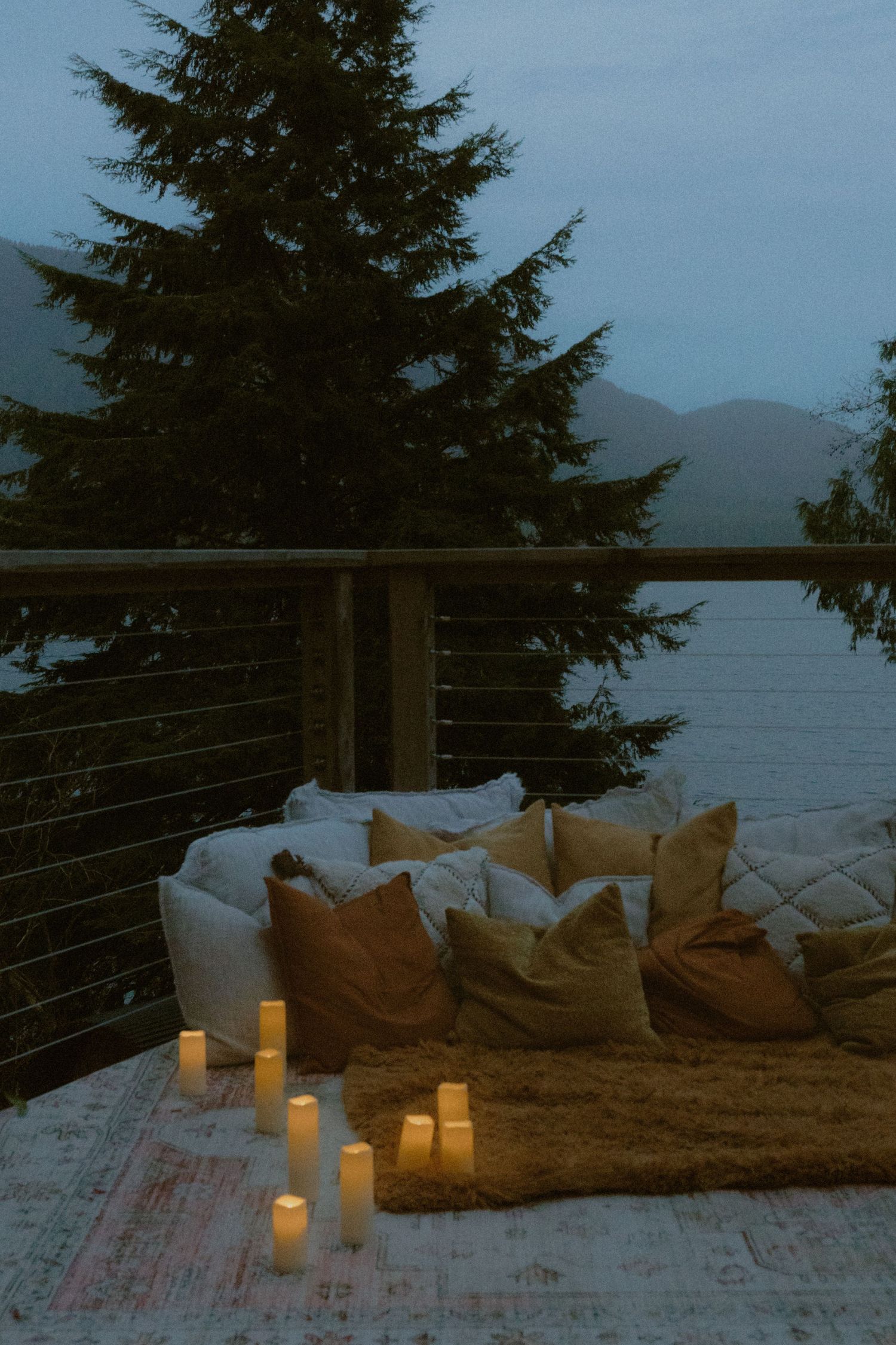 A cozy outdoor seating area with pillows and candles overlooking a lake at dusk.