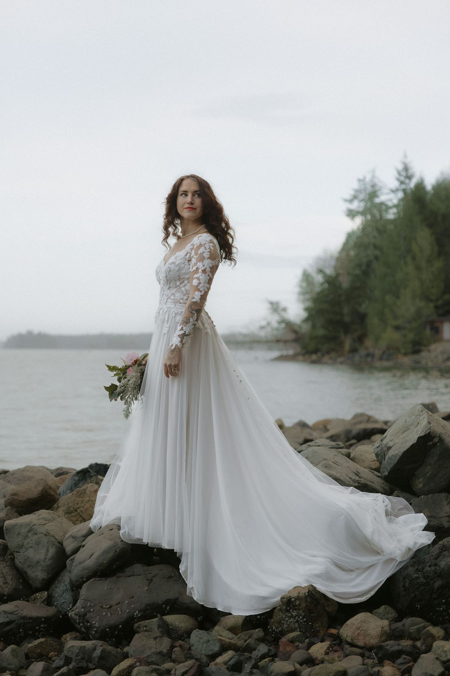 A flowing white wedding gown catches the breeze on a rocky beach with mountains in the background.