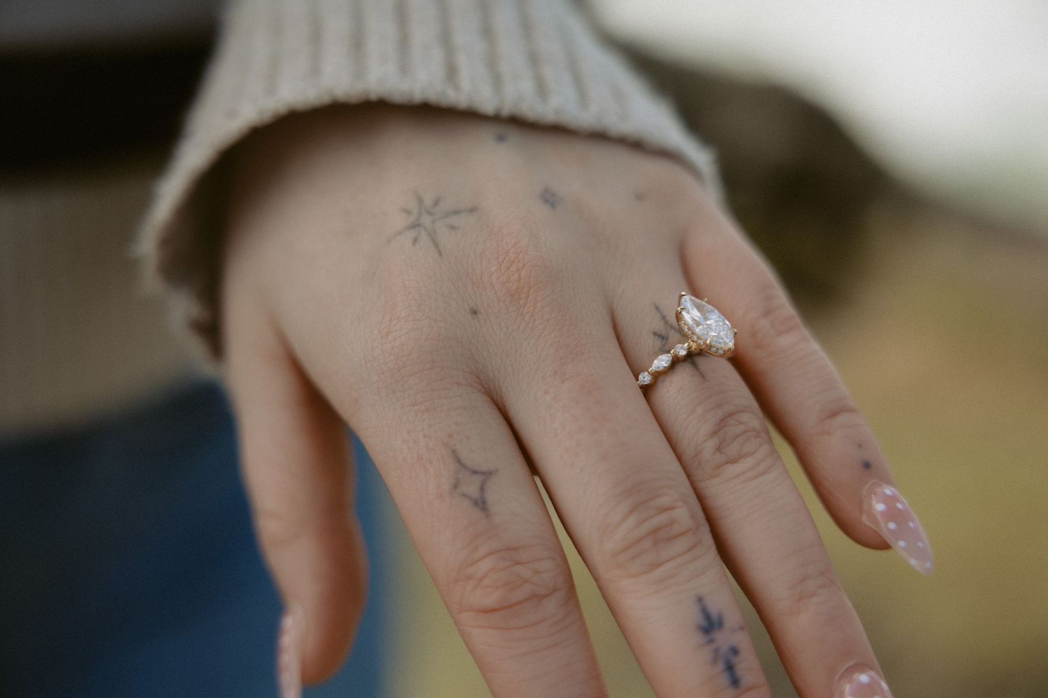 Close-up of tattooed hand wearing a pear-shaped diamond engagement ring with delicate band.