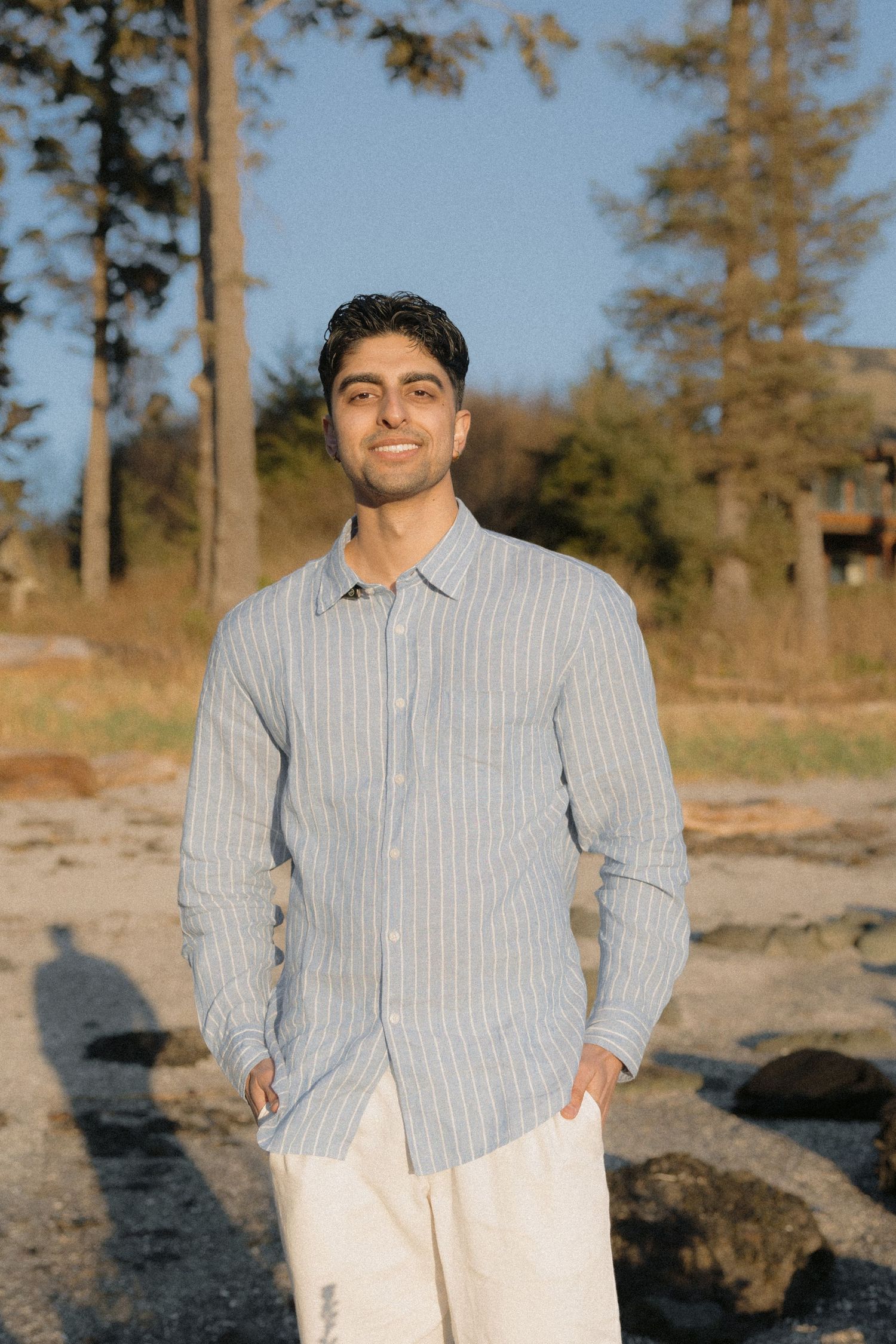 Smiling young man in striped blue linen shirt standing on rocky beach at golden hour.