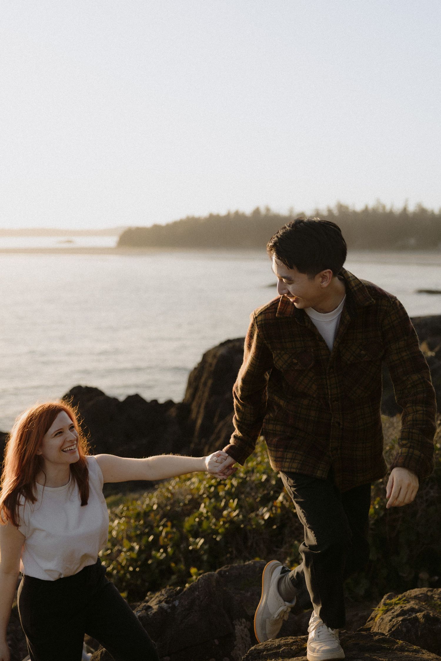 Couple holding hands on rocky coastal shore at sunset, woman with red hair laughing joyfully.