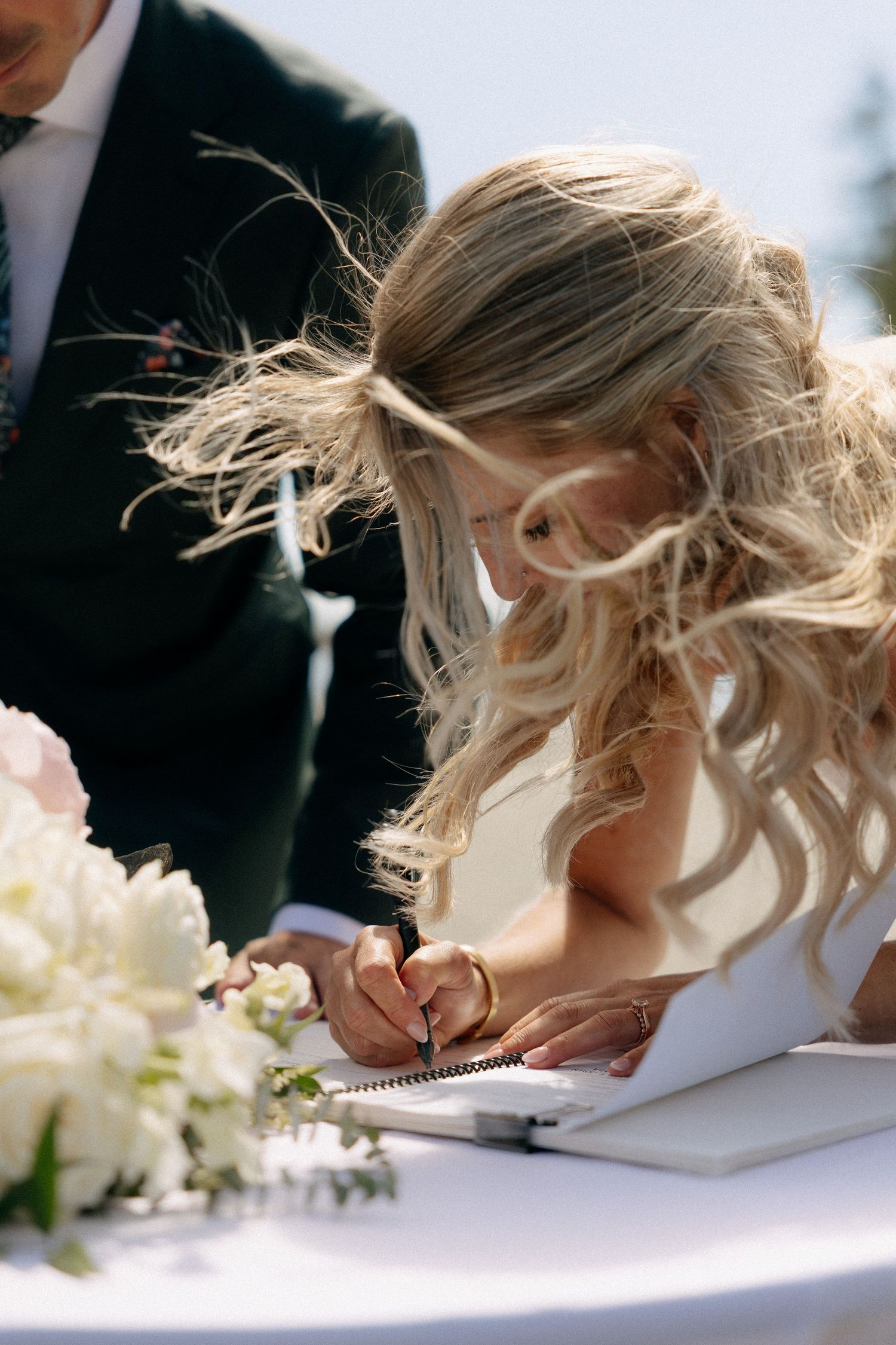 Bride with flowing blonde hair signing wedding registry at outdoor ceremony table with white floral arrangement.