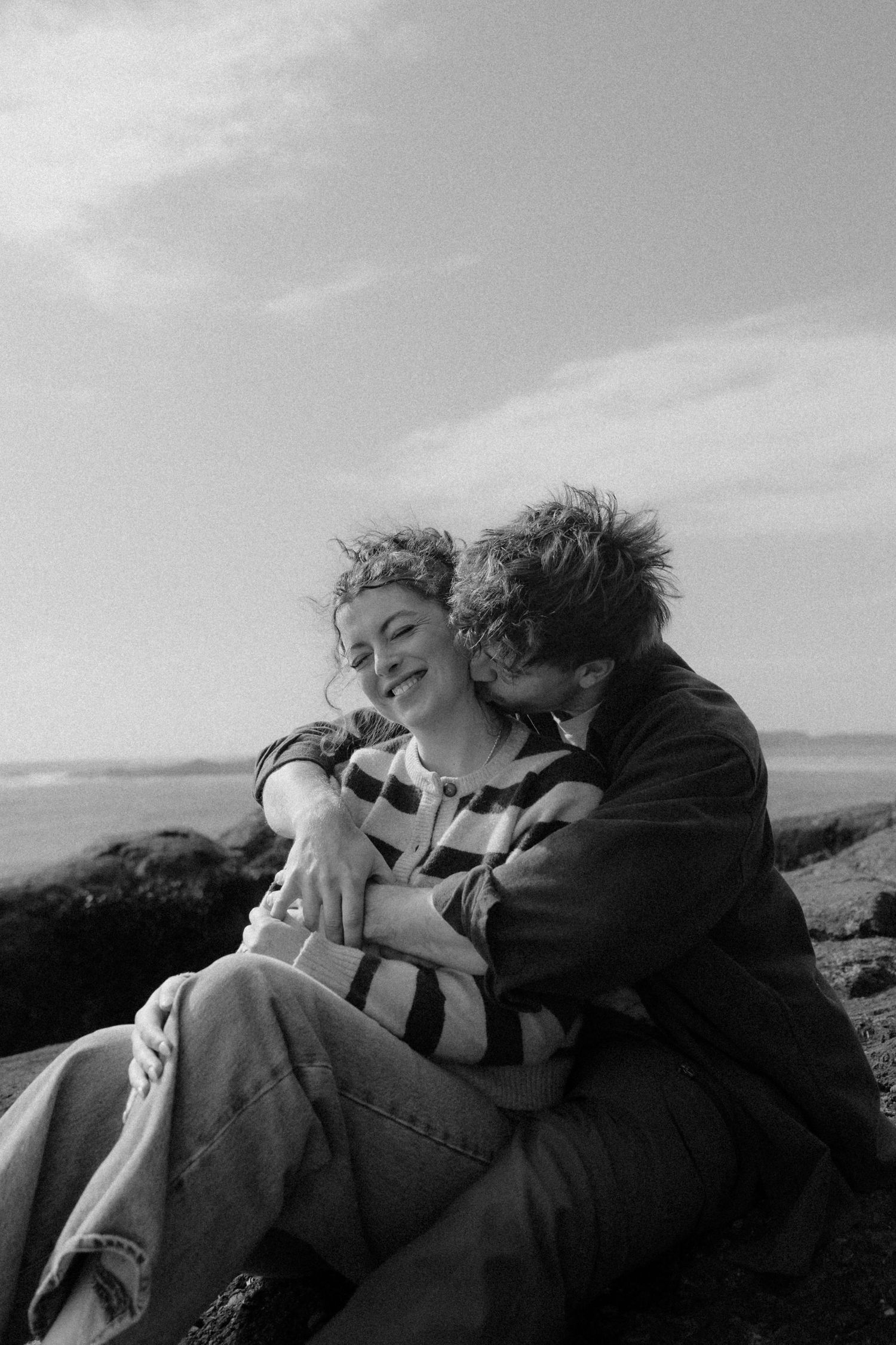 Black and white photo of couple cuddling and laughing together on rocky beach shoreline.