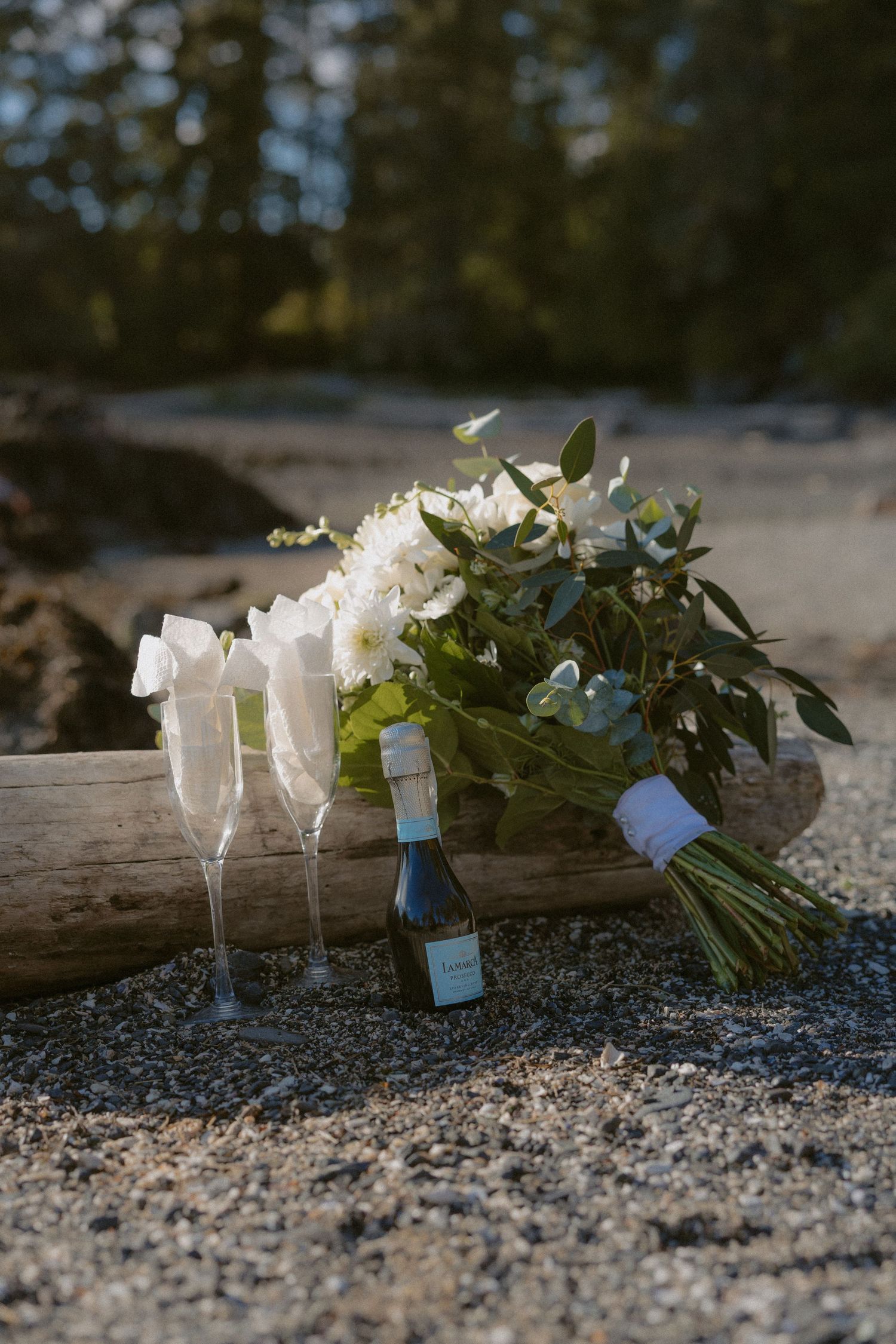 White wedding bouquet with champagne flutes and mini bottle on sandy beach at golden hour.