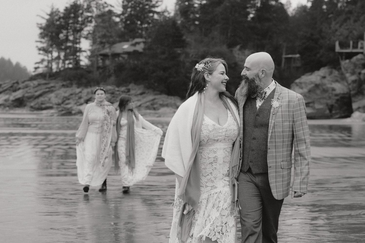 A couple shares an intimate moment on a beach while two figures in white walk behind them on the shore.